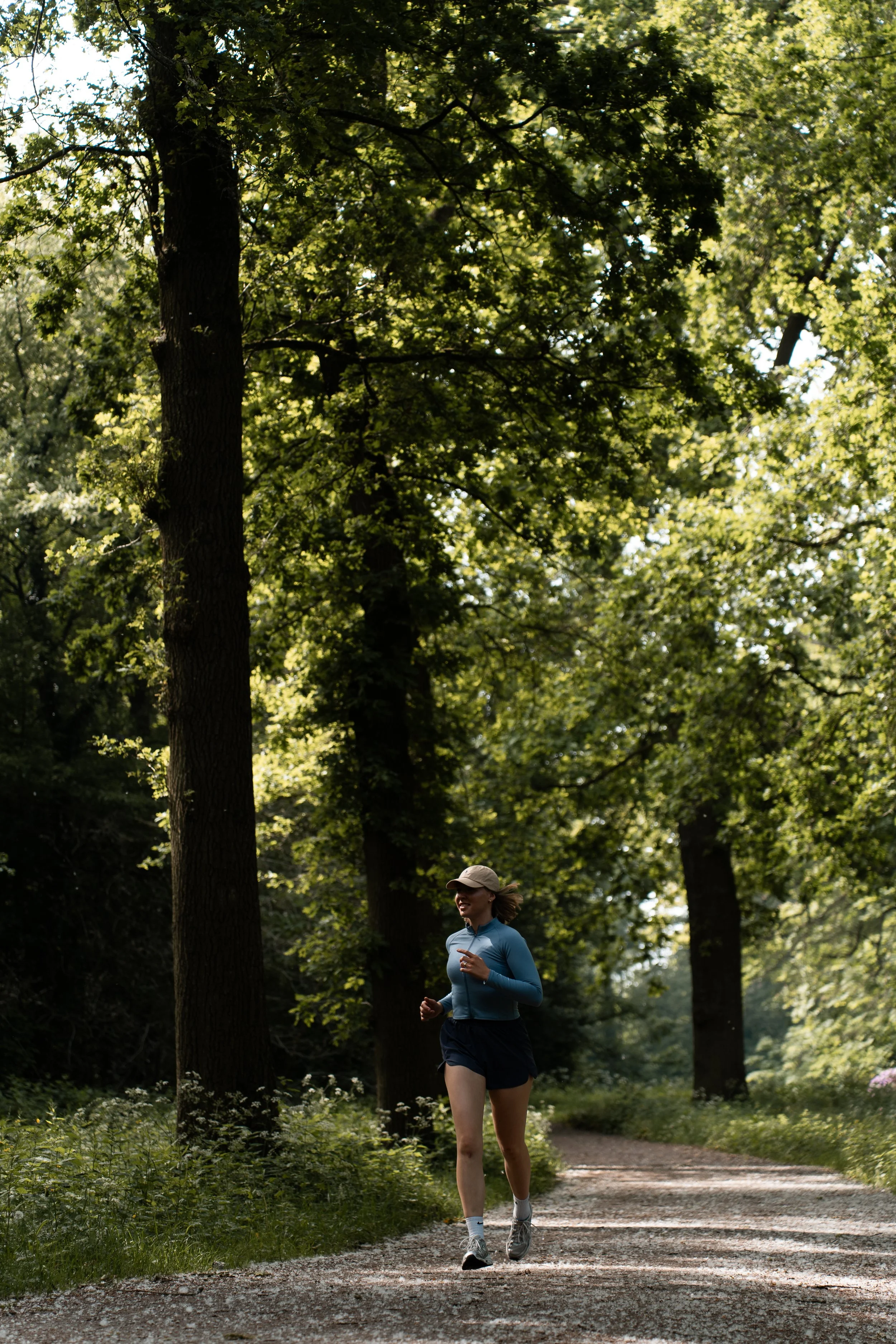 Een vrouw joggt op een bospad omgeven door groene bomen en struiken, gekleed in sportkleding en een pet.