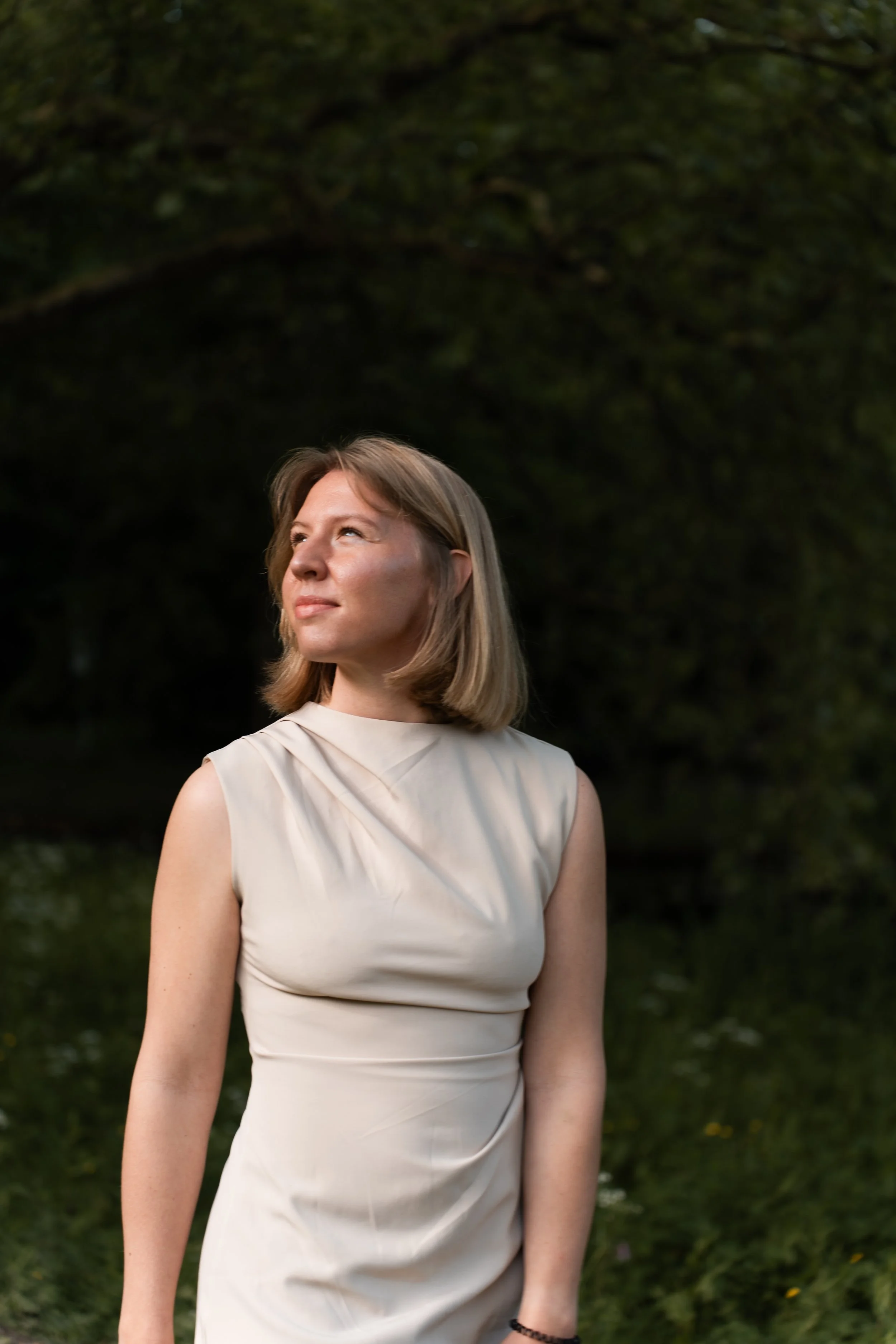 Portret van een vrouw met halflang blond haar die naar links kijkt, zittend in een park met groene bomen op de achtergrond, bij zonsondergang.