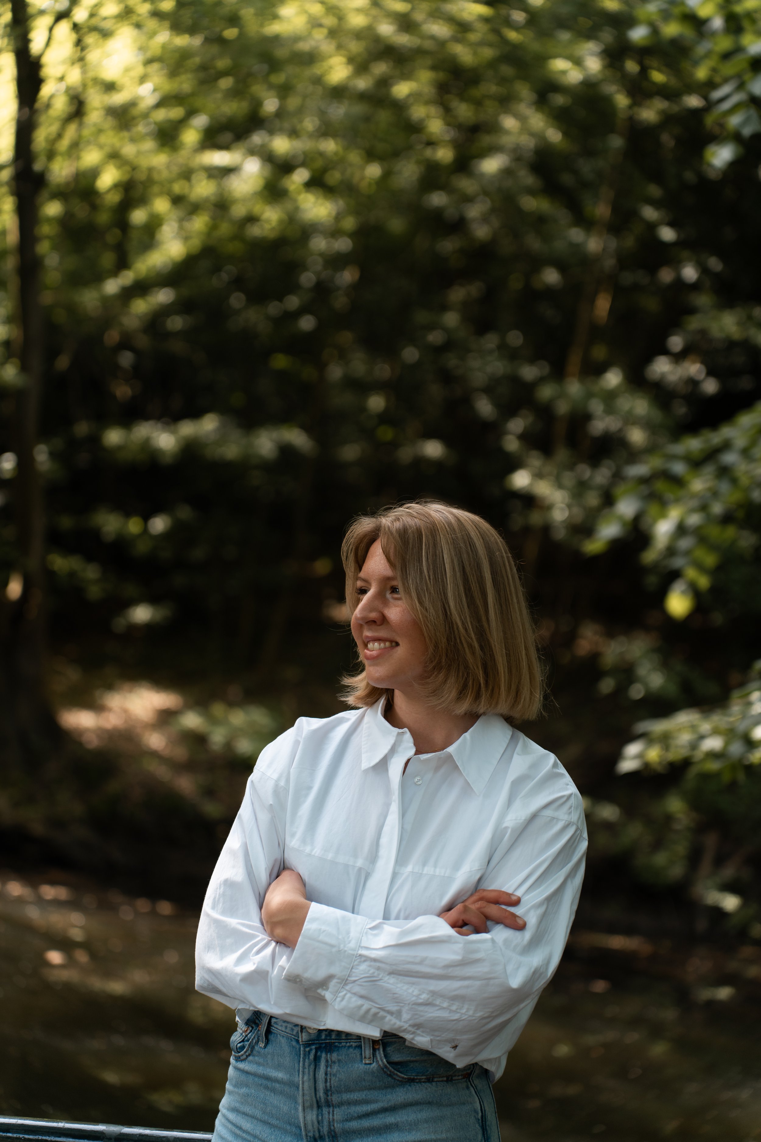 Portret van een vrouw met schouderlengde blond haar die binnen in een bos staat, gekleed in een witte blouse en blauwe spijkerbroek, lachend en met haar armen over elkaar.