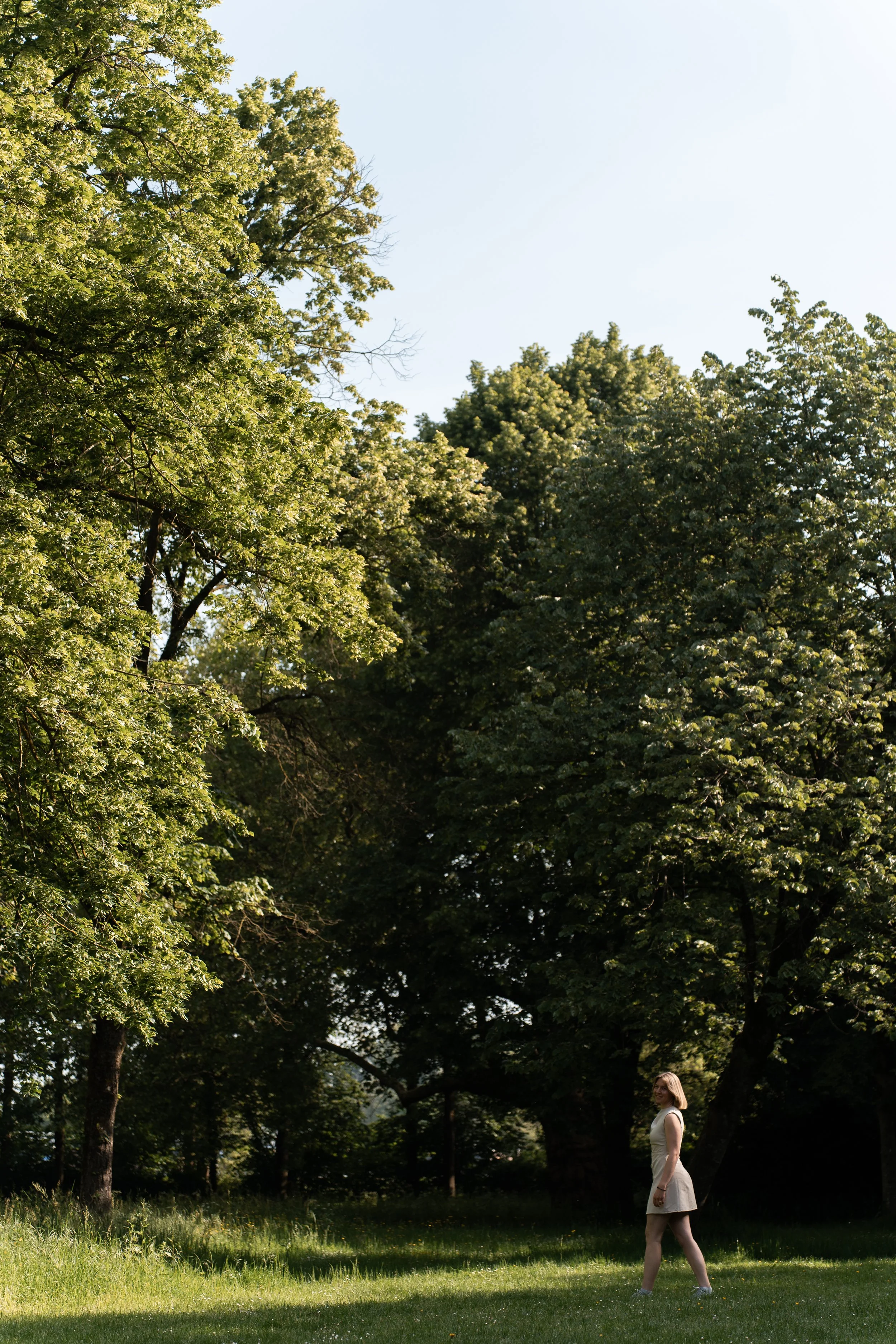 Een vrouw in een witte jurk wandelt door een groen park met grote bomen op een zonnige dag.