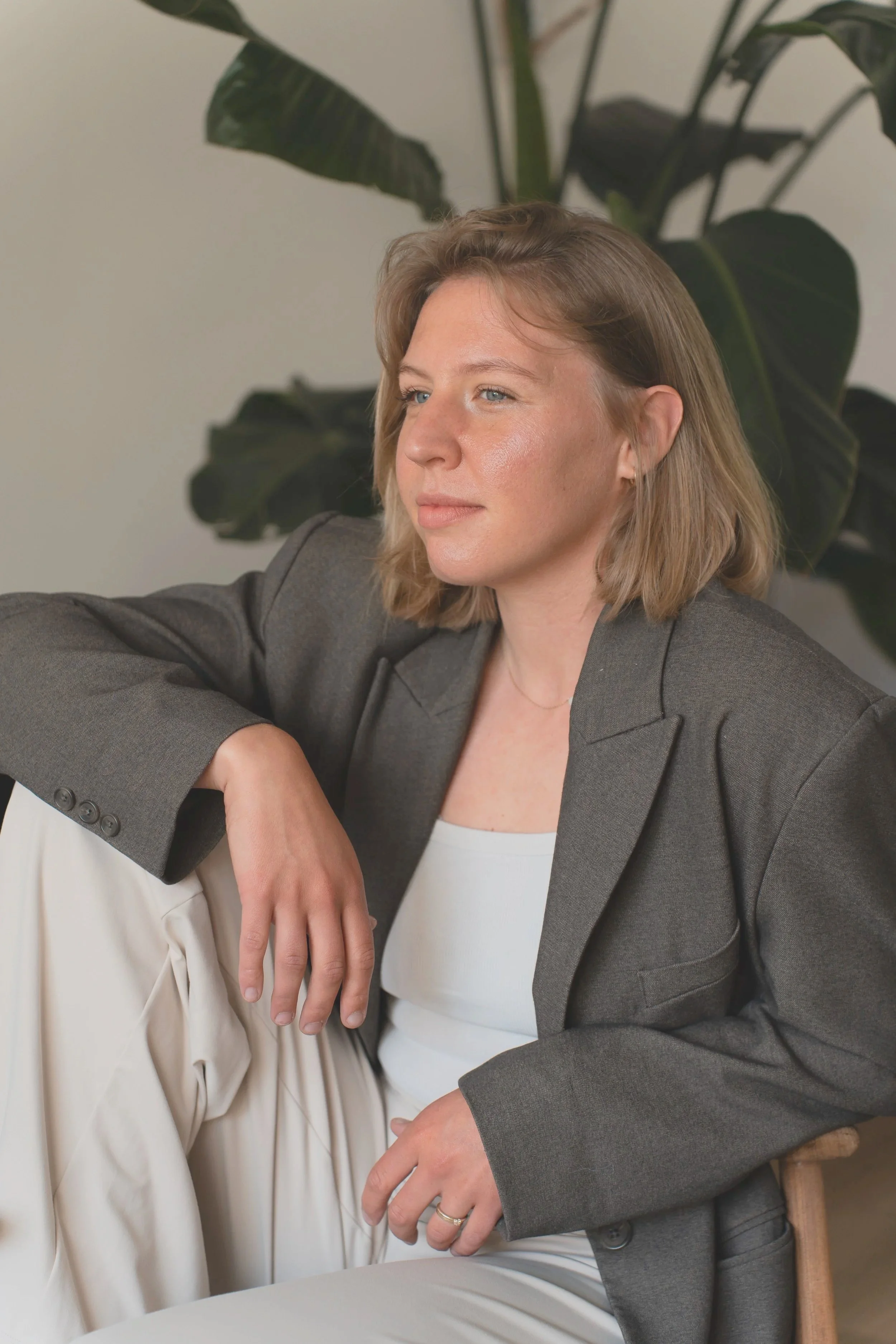 Een vrouw met schouderlang blond haar, gekleed in een grijze blazer en witte top, zit naast iemand anders, die een witte broek draagt. Ze zitten in een kamer met een grote groene plant op de achtergrond.