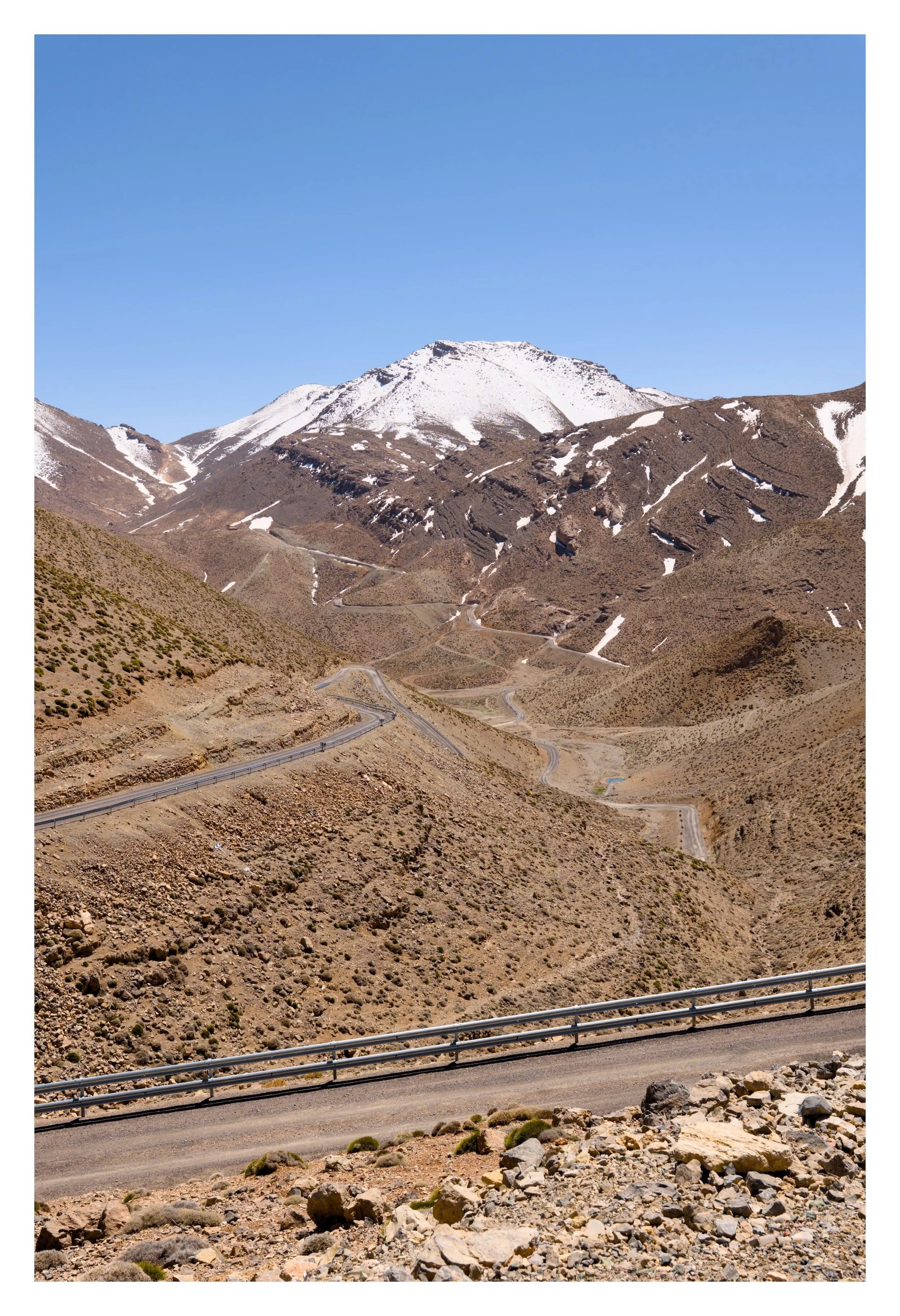 A winding mountain road with metal guardrails, leading through a rocky, arid landscape with sparse vegetation. In the background, snow-capped mountains under a clear blue sky.