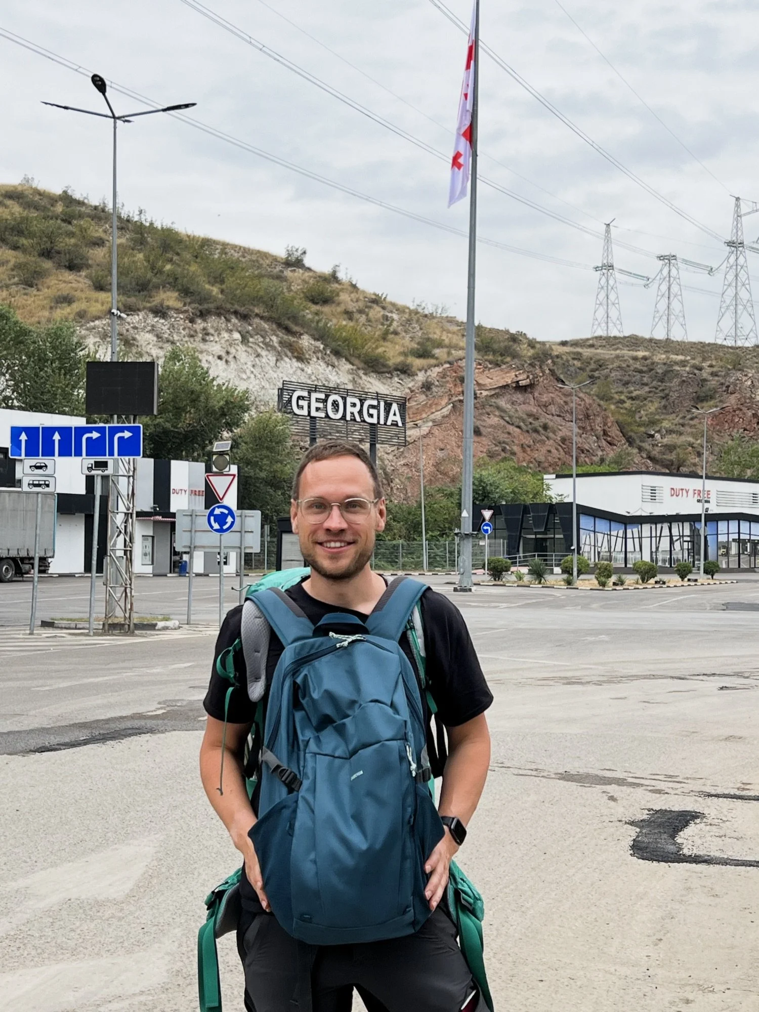 A smiling man wearing glasses, a black t-shirt, and a teal backpack standing in a parking lot with a Georgia sign in the background, hills, power lines, and a cloudy sky.