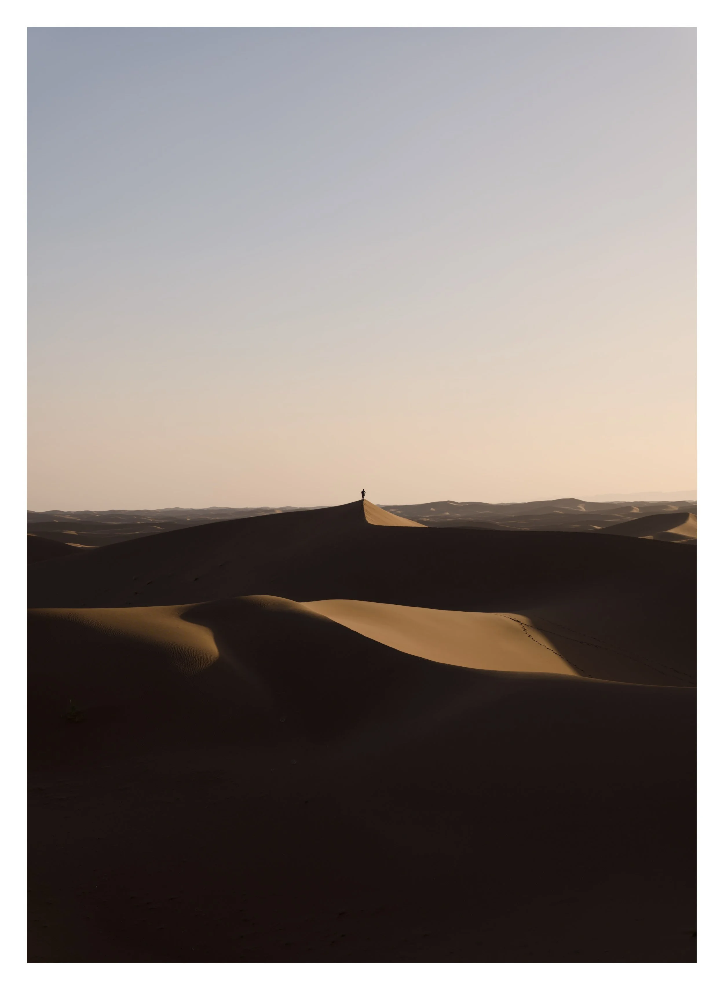 A person standing on top of a sand dune in a vast desert landscape during sunset or sunrise.