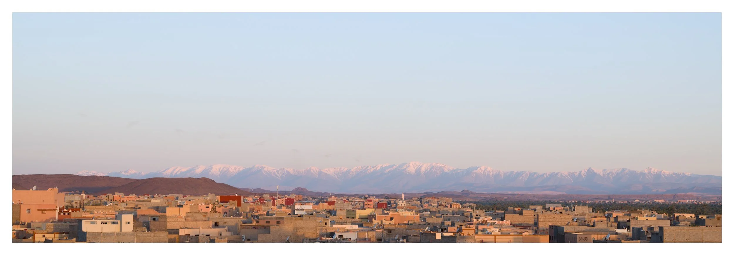 Panoramic view of a desert city with flat rooftop buildings, distant snow-capped mountains under a clear sky.