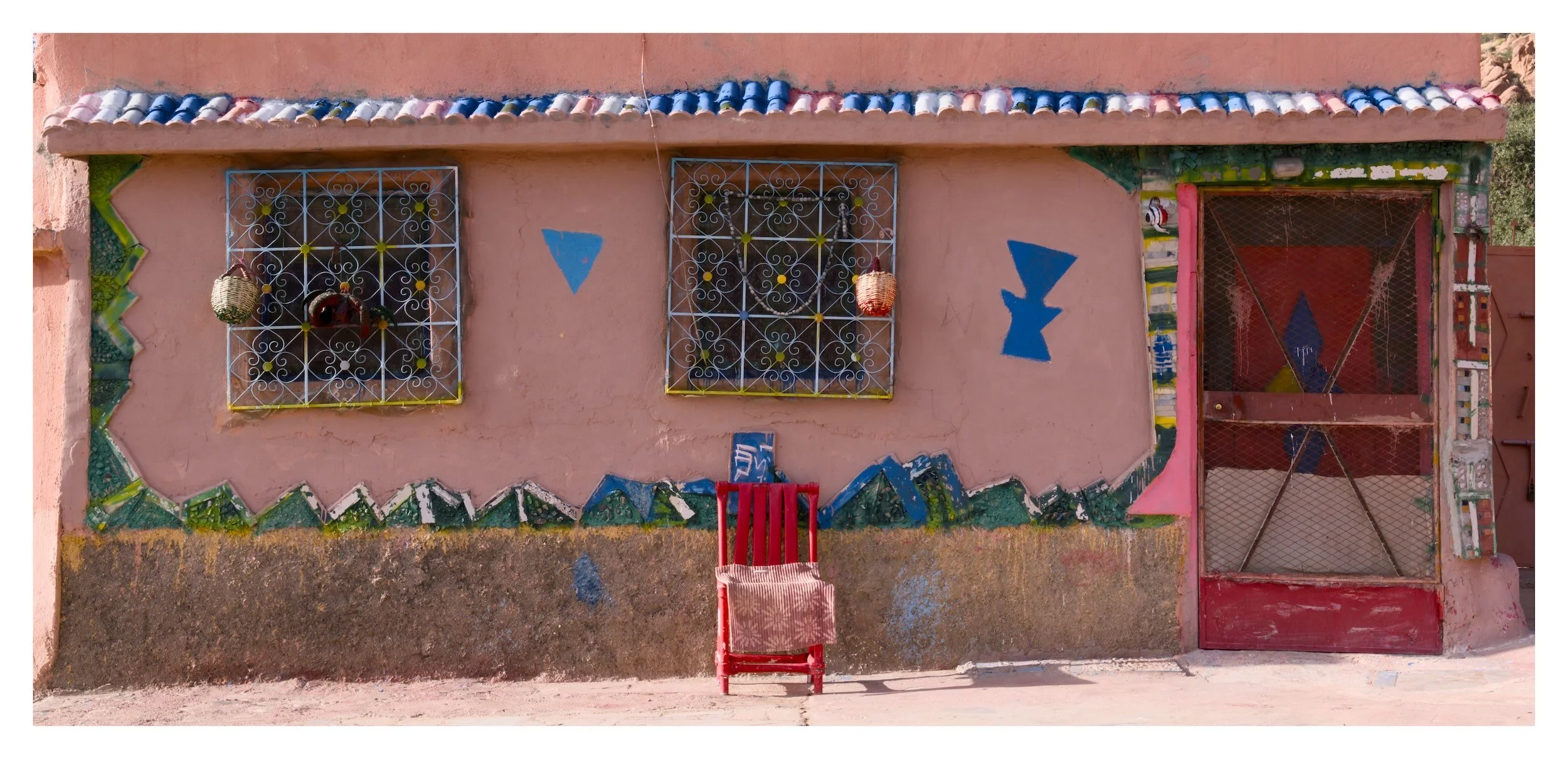 A pink wall with blue and yellow decorative elements and two barred windows. Baskets hang from the wall. A red chair with a striped cloth is positioned in front of the wall, and a pink metal gate is on the right side of the wall.