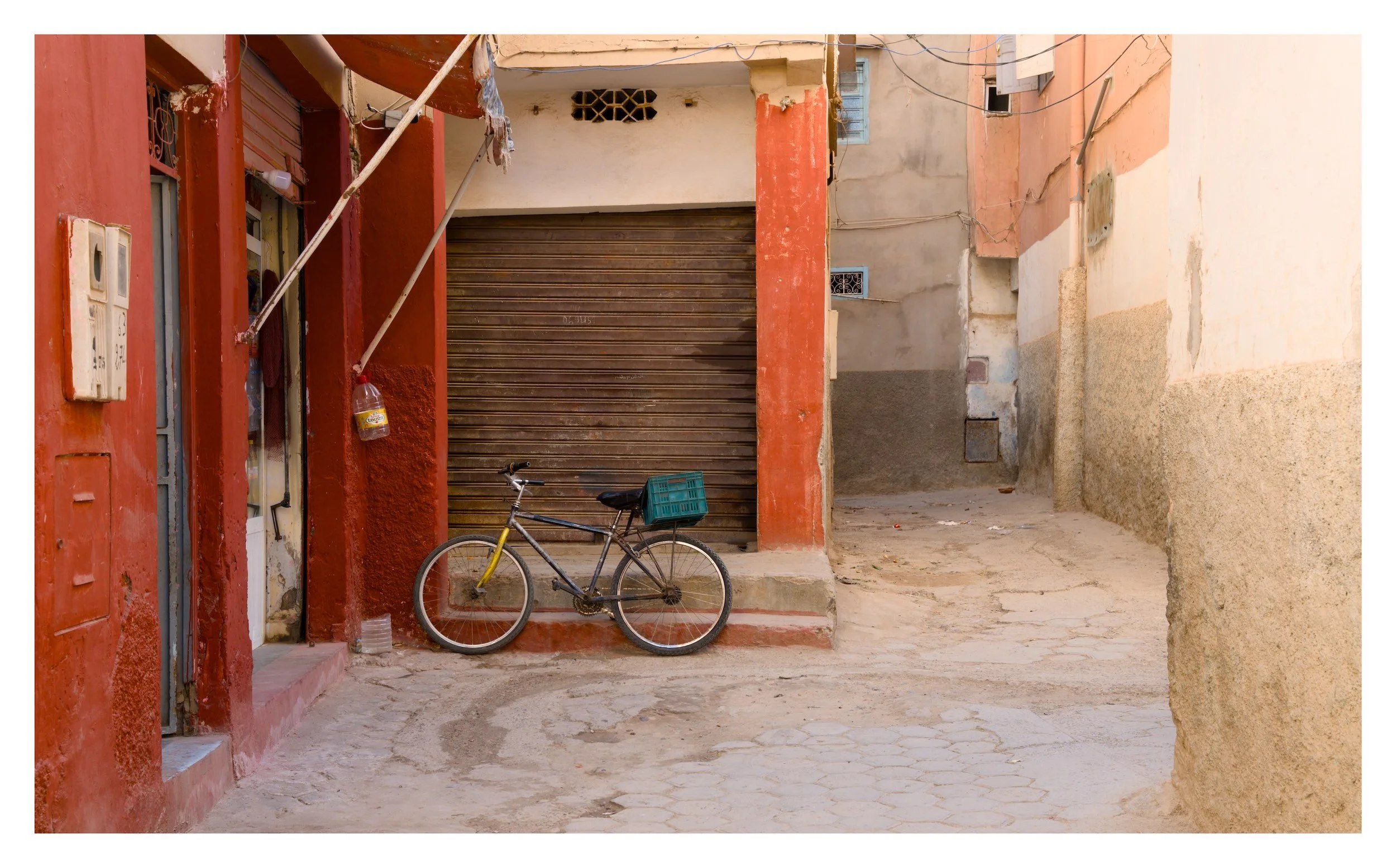 A narrow street with a closed metal roll-up door, a bicycle with a green crate attached to the back, and residential buildings painted in red, beige, and cream. The street has a hexagonal patterned concrete surface.