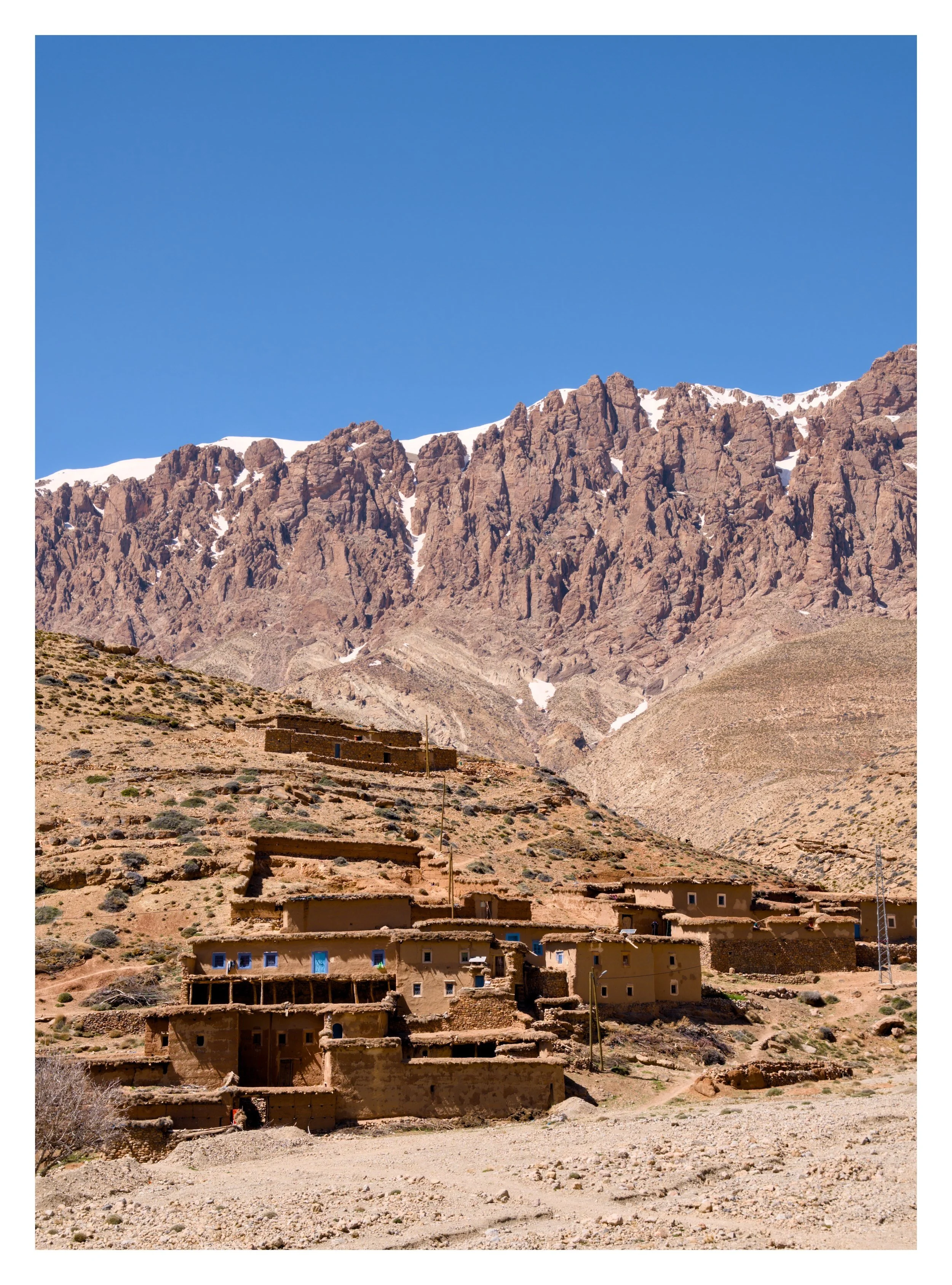 Mountain village with traditional adobe houses at the base of tall rocky mountains with snow patches under a clear blue sky.