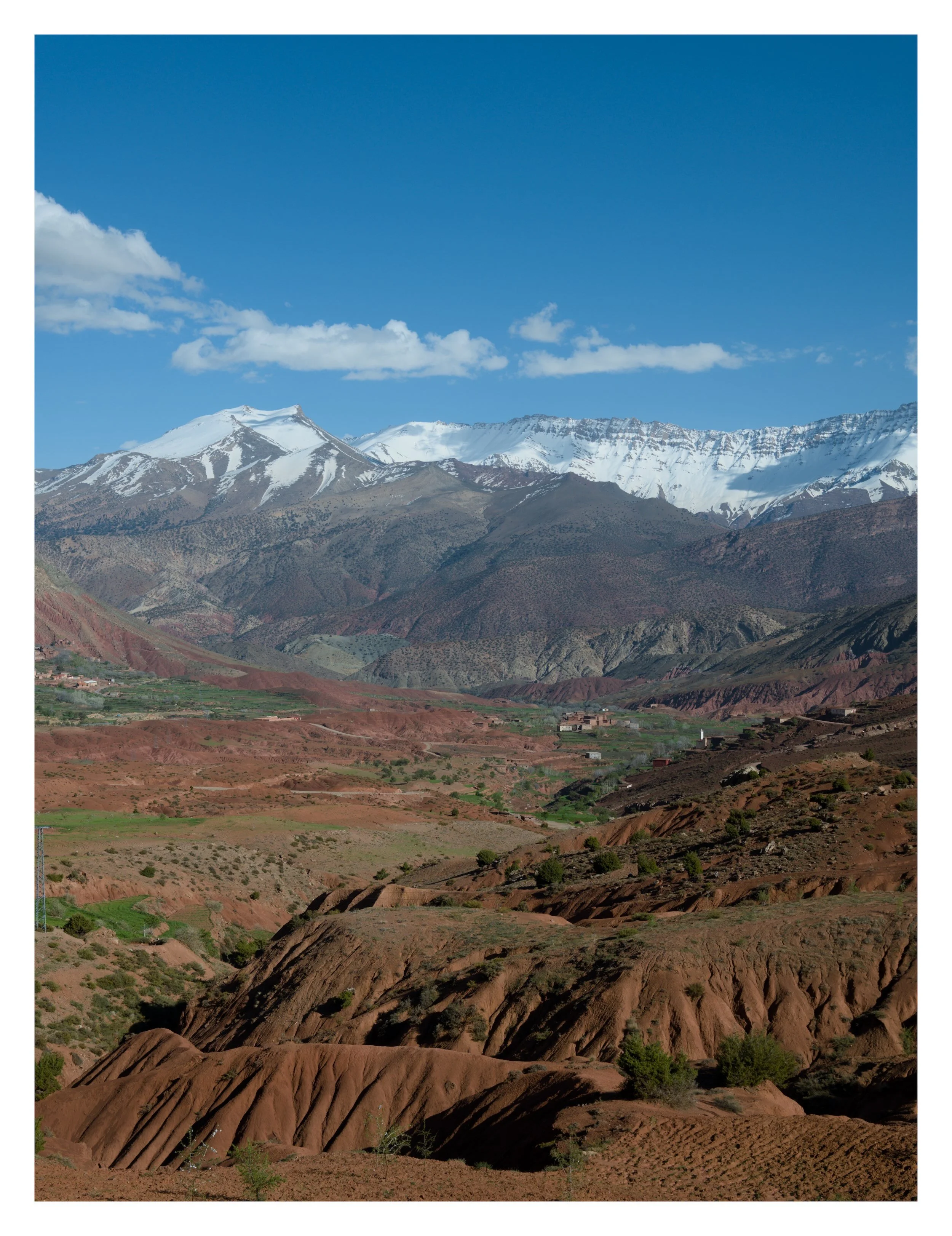 Scenic mountain landscape with snow-capped peaks, rugged terrain, and sparse vegetation under a blue sky with scattered clouds.