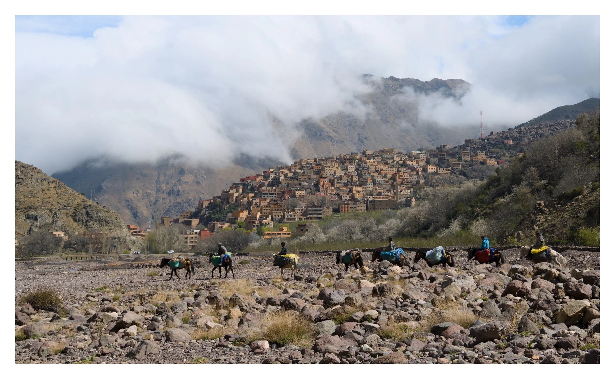 A mountainous landscape with a village on the hillside, clouds covering the mountain peaks, and a group of people and donkeys in the foreground, likely in a rural area.