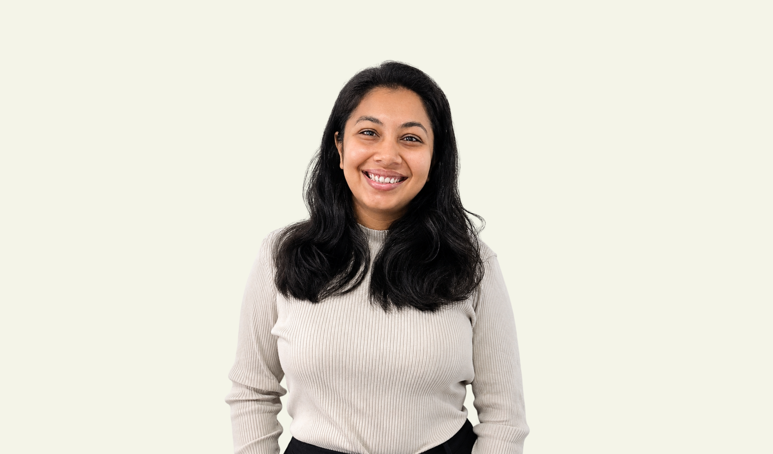 A woman with long black wavy hair smiling, wearing a beige ribbed long sleeve top and black pants, standing against a plain light background.