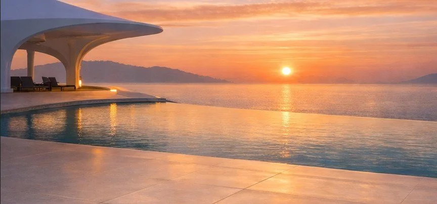 Sunset over an infinity pool with modern lounge chairs and a scenic view of mountains and water.
