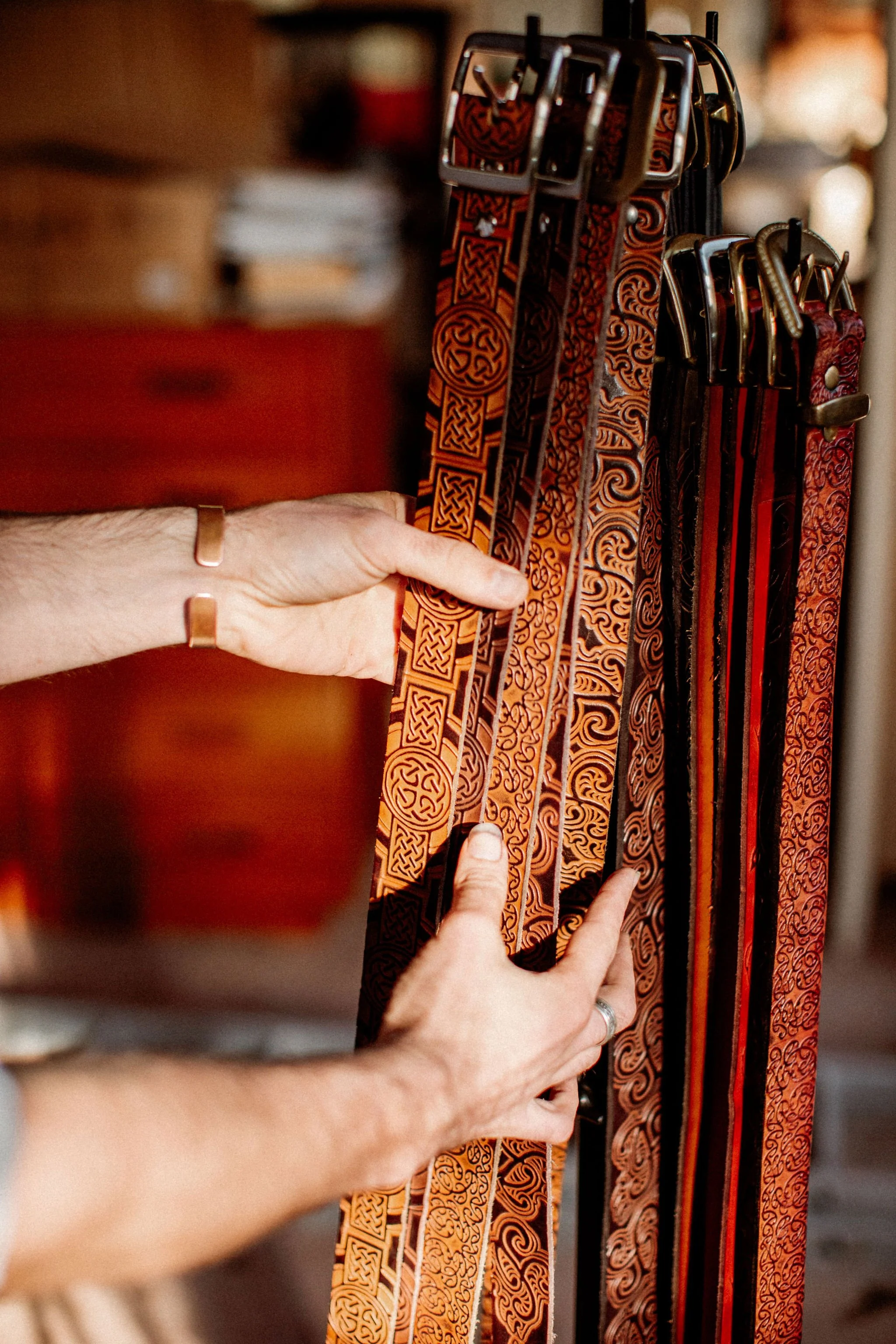 A person shopping for belts, holding several leather belts with intricate patterns in orange, red, and black colors at a store.