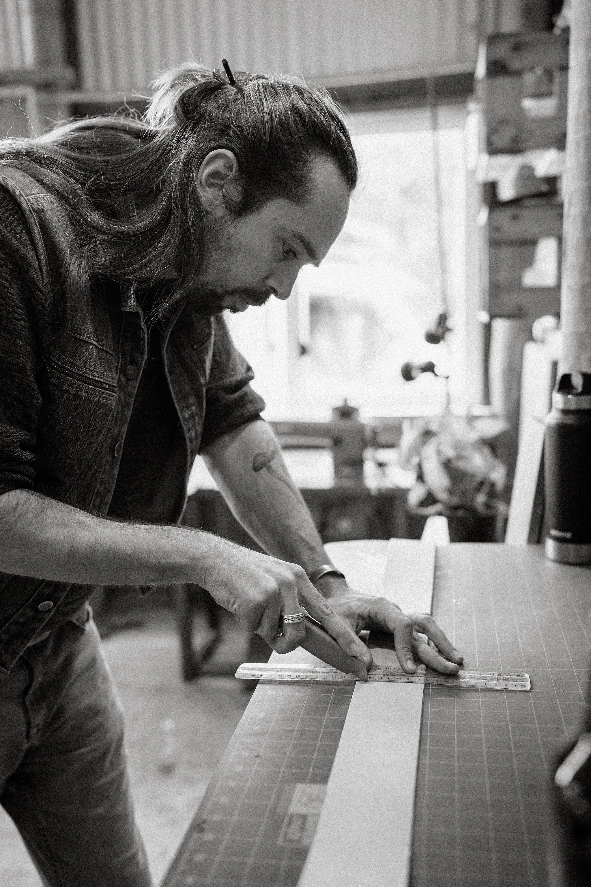 A man with long hair, beard, and tattoos is working on a craft project at a table, using a ruler and cutting mat in a workshop or studio.