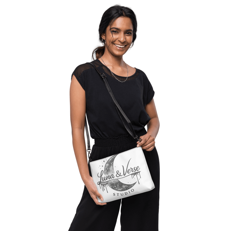 A young woman with dark hair and earrings smiling, wearing a black shirt and black pants, holding a white bag with a moon and star design and the words Luna & Verse Studio, standing against a black background.
