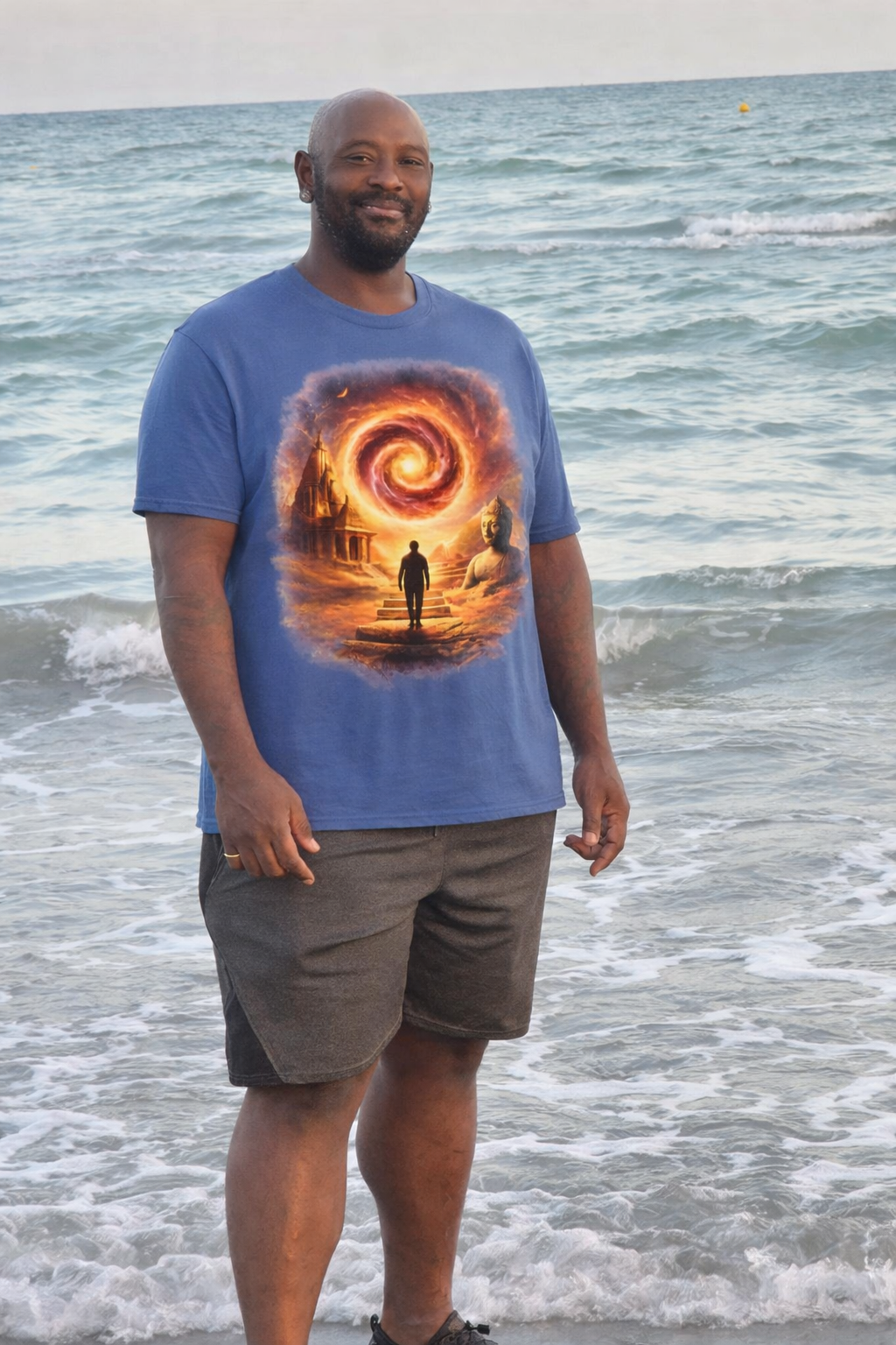 A man standing on the beach at the shoreline with the ocean waves behind him, wearing a blue T-shirt featuring a fantasy scene and smiling at the camera.
