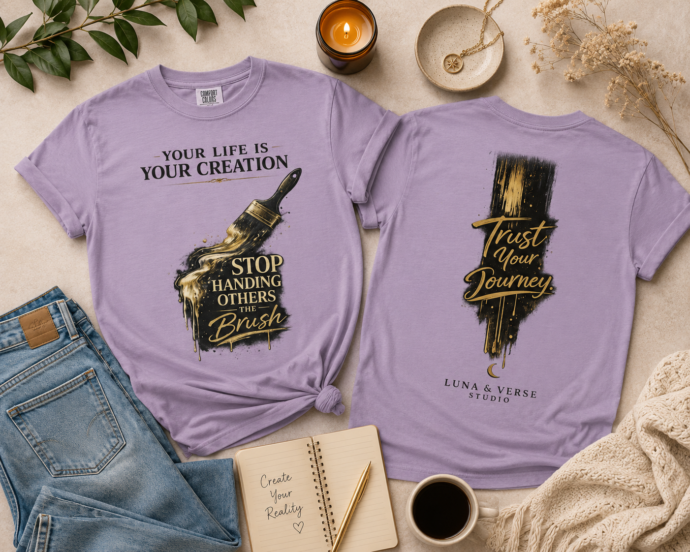 Two lavender T-shirts with motivational messages, one with a paintbrush and the other with a painted background, surrounded by a notebook, a cup of coffee, a candle, green leaves, dried flowers, and a cream-colored blanket.