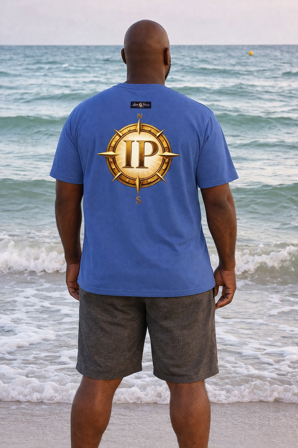 A man standing on the beach facing the ocean, wearing a blue shirt with a gold compass emblem and the letters 'IP' in the center.