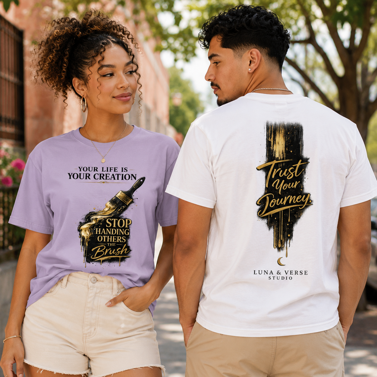 A young woman and man are standing outdoors on a sunny day, wearing t-shirts with inspiring messages. The woman has curly hair and is wearing a lavender shirt with gold and black graphics and text reading, 'Your life is your creation. Stop handing others the brush.' The man has short dark hair and is wearing a white shirt with black and gold graphics and text saying, 'Trust your journey,' with smaller text 'Luna & Verse Studio' underneath.