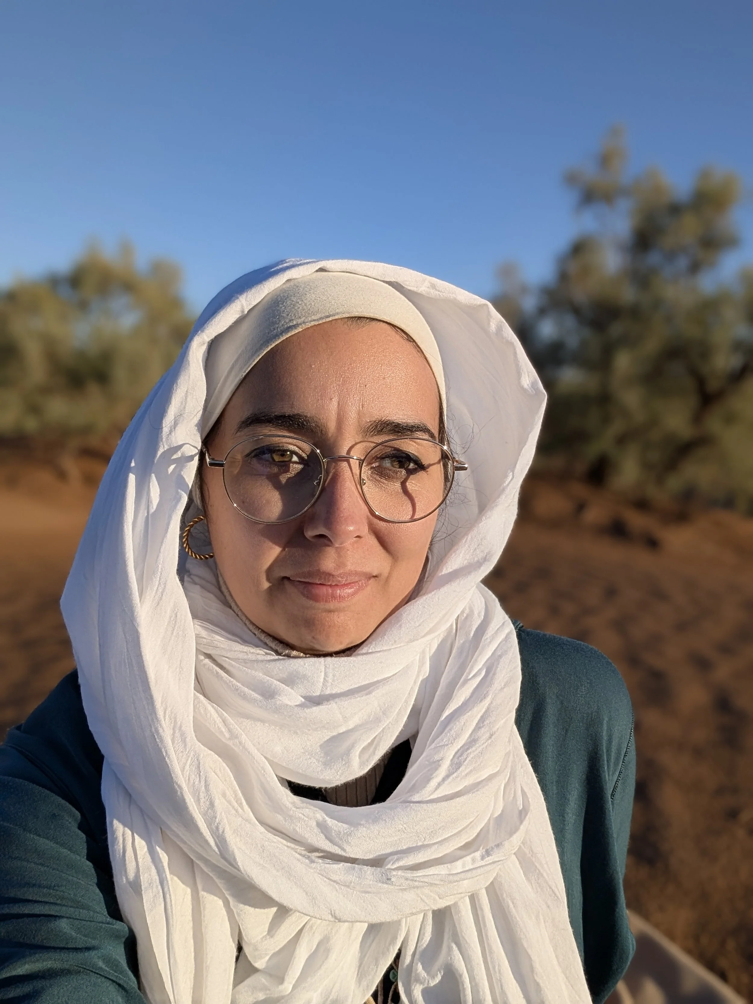 Portrait d'une femme portant des lunettes portant un chèche blanc, dans le désert sous un ciel bleu, avec des arbres en arrière-plan, probablement lors d'une journée ensoleillée.