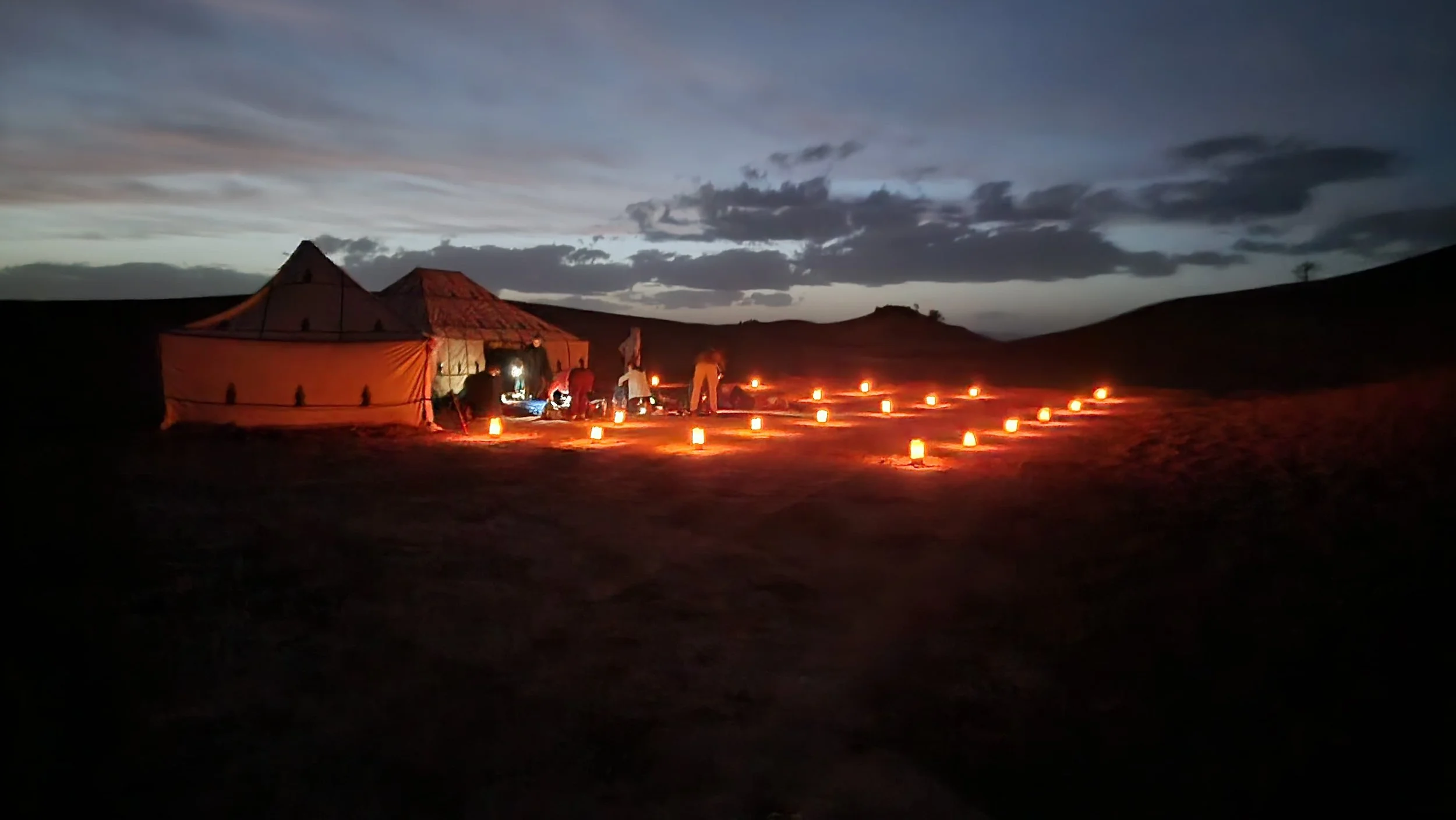 Tente beige avec personnes à l'extérieur durant la nuit, éclairée par des lanternes orangées dans un paysage désertique avec des dunes en arrière-plan.