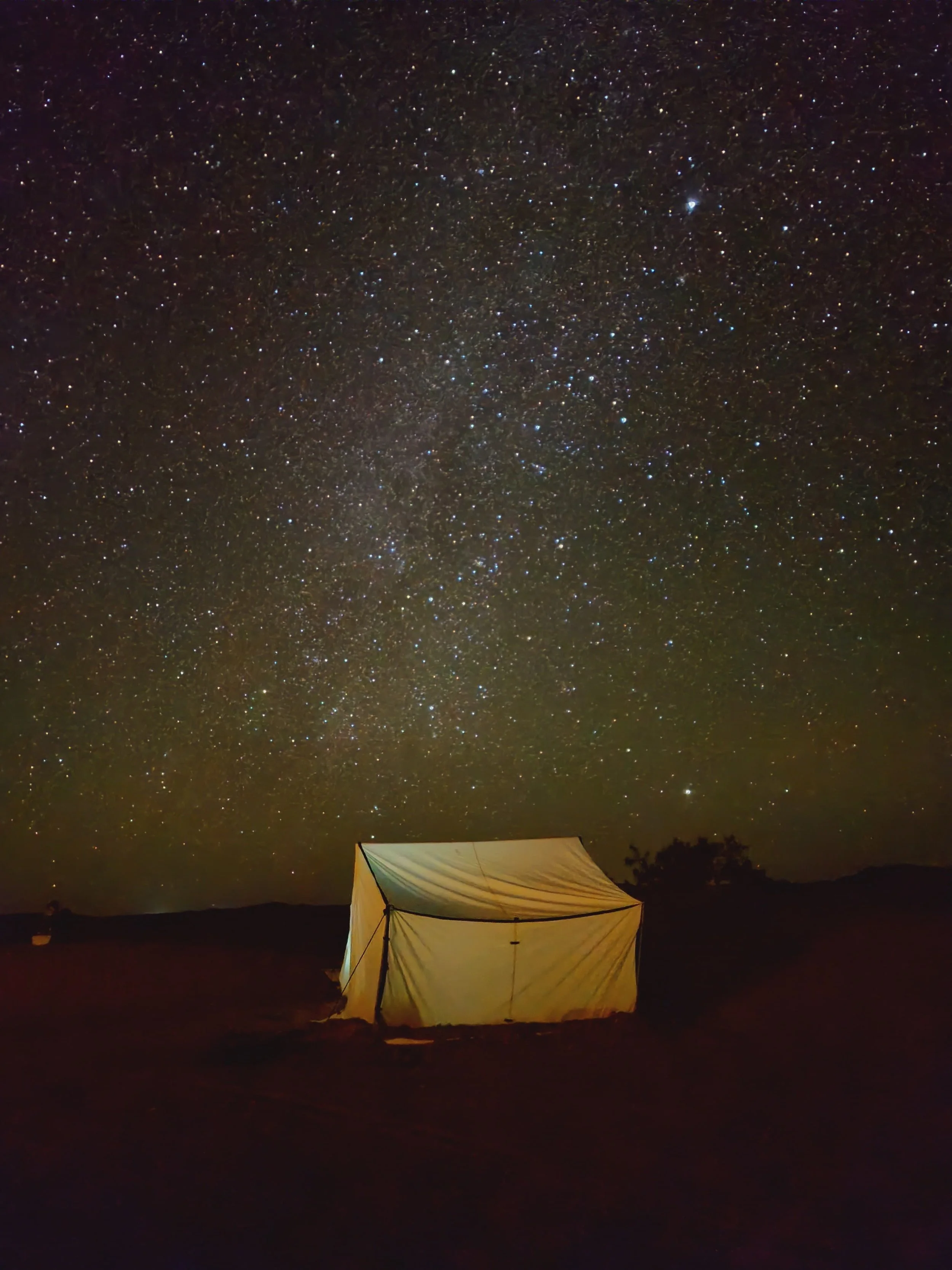 Ciel étoilé avec de nombreuses étoiles brillantes au-dessus d'une tente éclairée au sol, dans un paysage nocturne désertique ou montagneux.