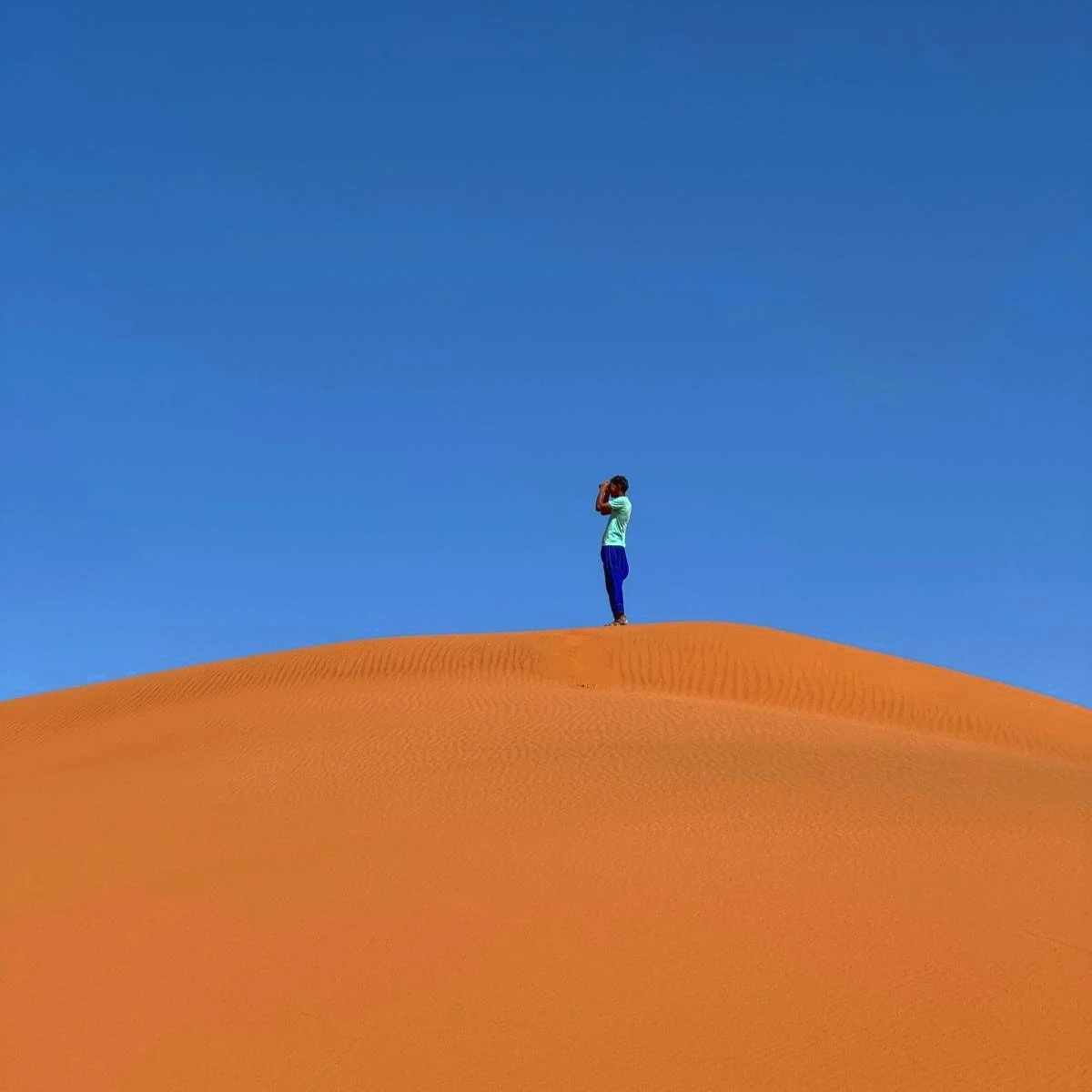 Personne debout sur une dune de sable orange dans un désert, regardant à travers des jumelles, ciel bleu clair en arrière-plan.