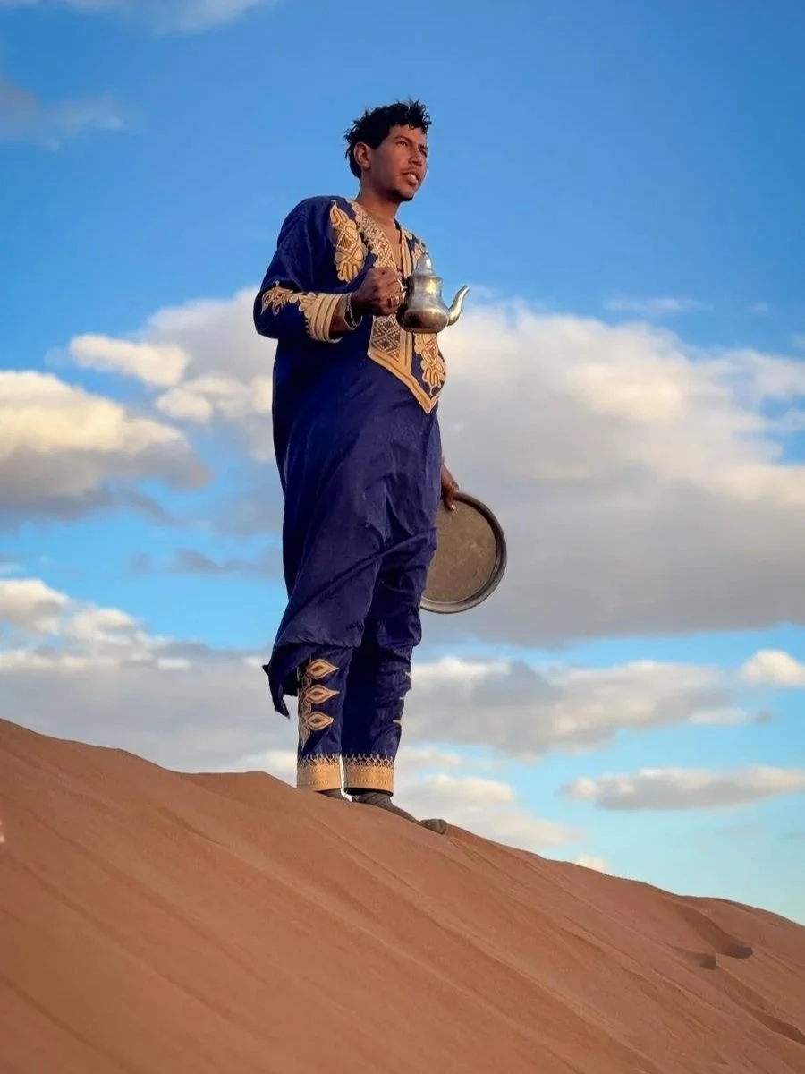 Un nomade marocain debout sur une dune de sable dans le désert, portant une tenue traditionnelle bleue avec des motifs dorés, tenant une théière en métal dans une main et un plateau dans l'autre, avec un ciel bleu et des nuages en arrière-plan.