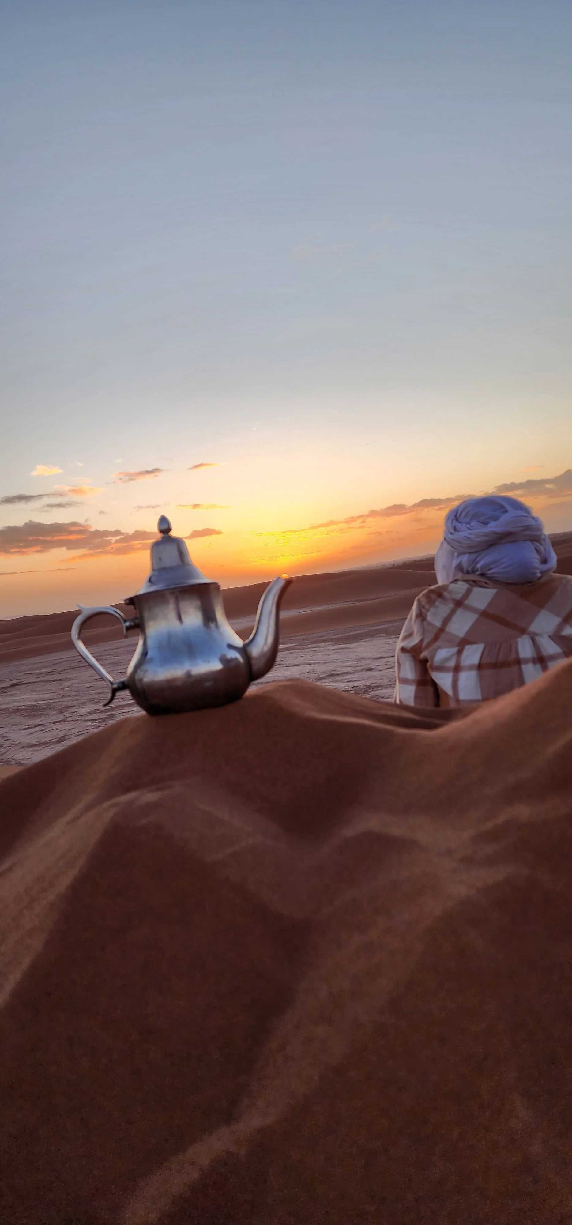 Une femme portant un turban blanc regarde un coucher de soleil dans le désert, avec une théière en métal posée sur le sable.