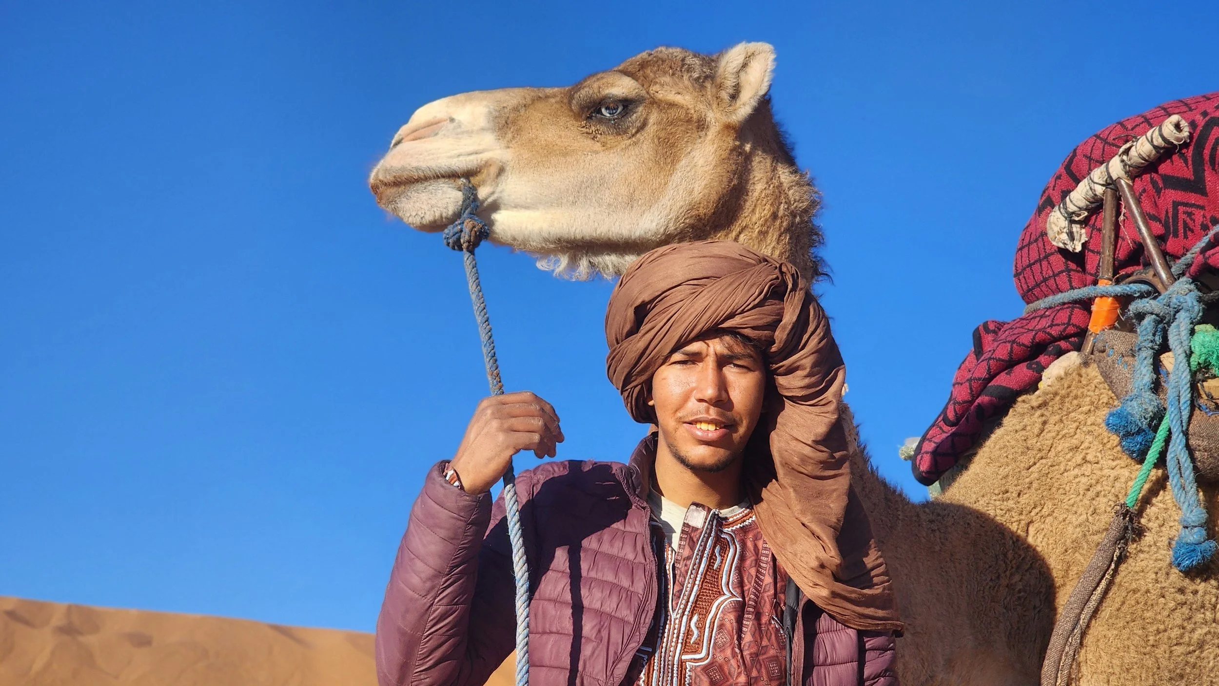 Un nomade marocain portant un turban se tient à côté d'un dromadaire dans un désert, sous un ciel bleu.