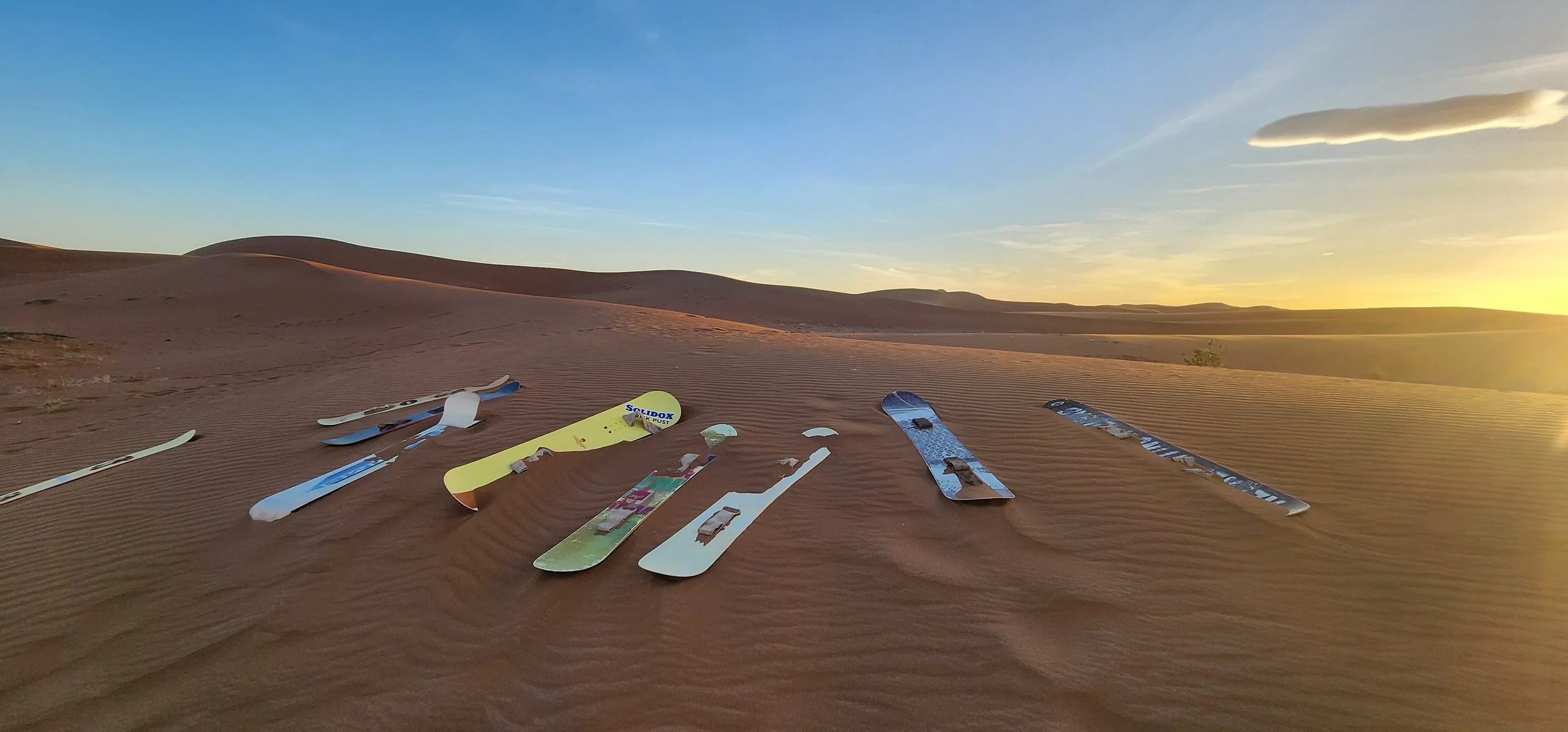 Dans le désert, plusieurs planches de sandboard reposent sur le sable fin, avec des dunes en arrière-plan et un ciel clair au coucher du soleil.