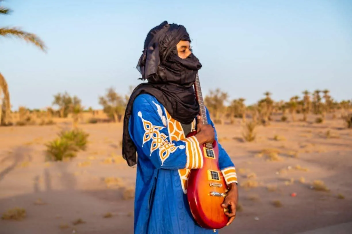 Un musicien nomade tenant une guitare électrique dans le désert marocain avec quelques arbres lointains.