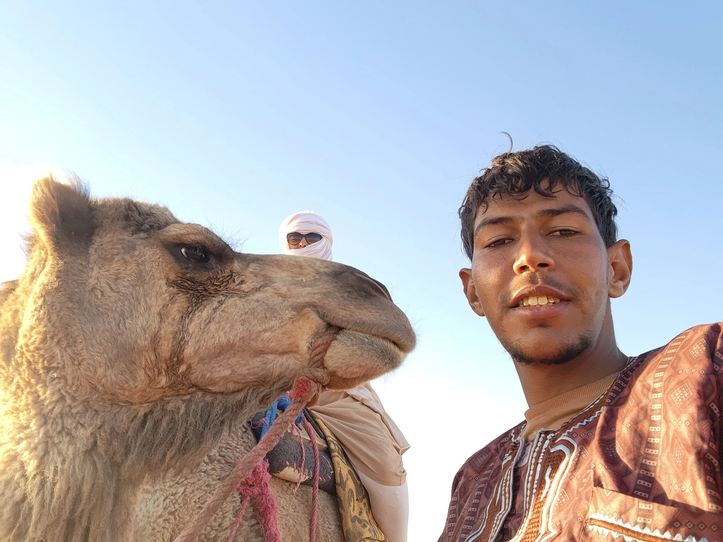 Un nomade et une touriste prennent un selfie avec un dromadaire dans un ciel clair.