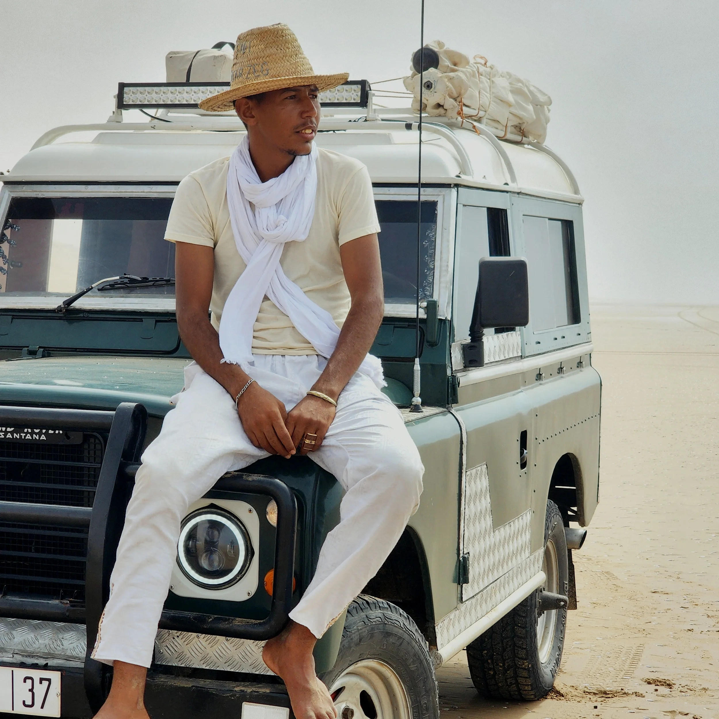 Un nomade marocain assis sur un 4x4, portant un chapeau en paille et un chèche blanc, avec un paysage désertique en arrière-plan.