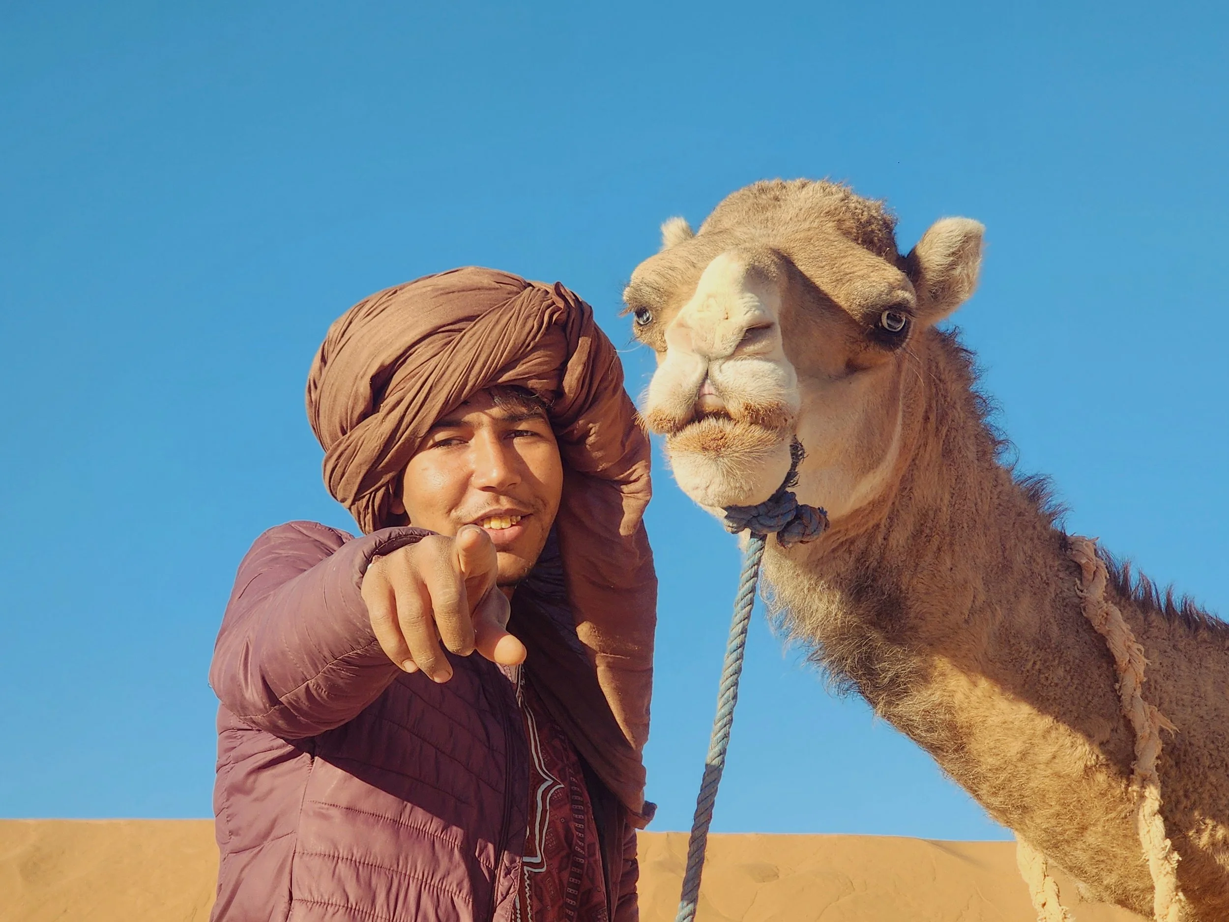 Un nomade du désert souriant avec un turban marron et une veste violette, pointant vers la caméra, se tenant à côté d’un chameau dans le désert sous un ciel bleu clair.