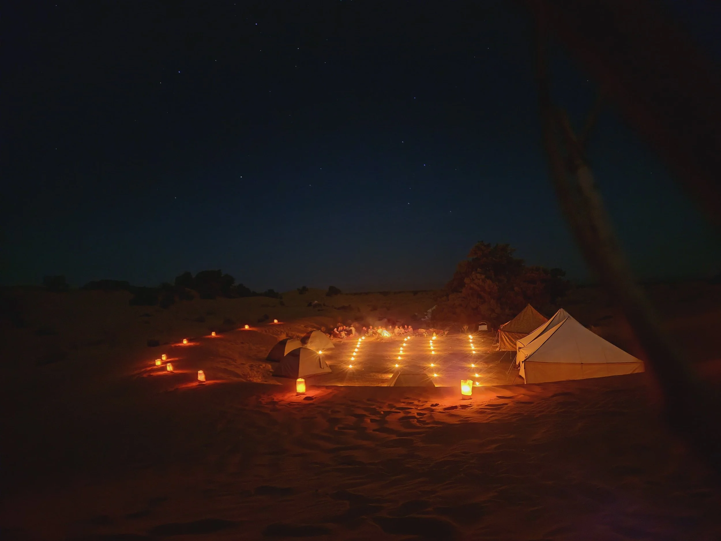 Campsite illuminé par des lanternes dans un paysage nocturne avec un ciel étoilé et des tentes, arbres et collines en arrière-plan.