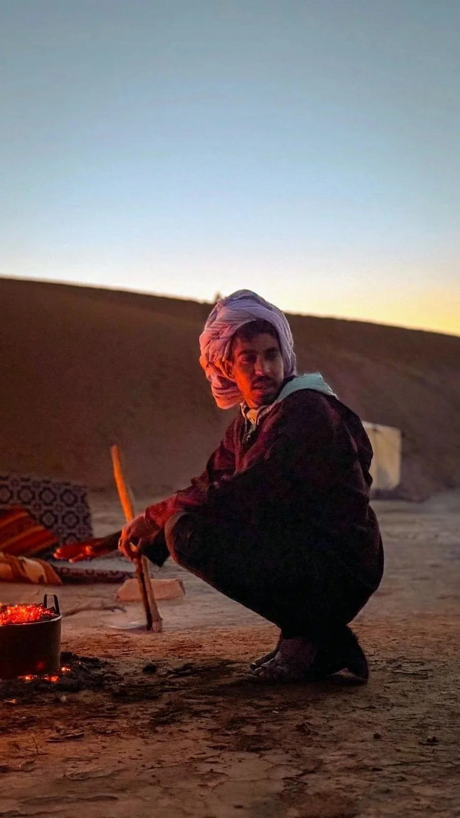 Un nomade du Maroc, accroupi près d'un feu de camp dans le désert au coucher du soleil.