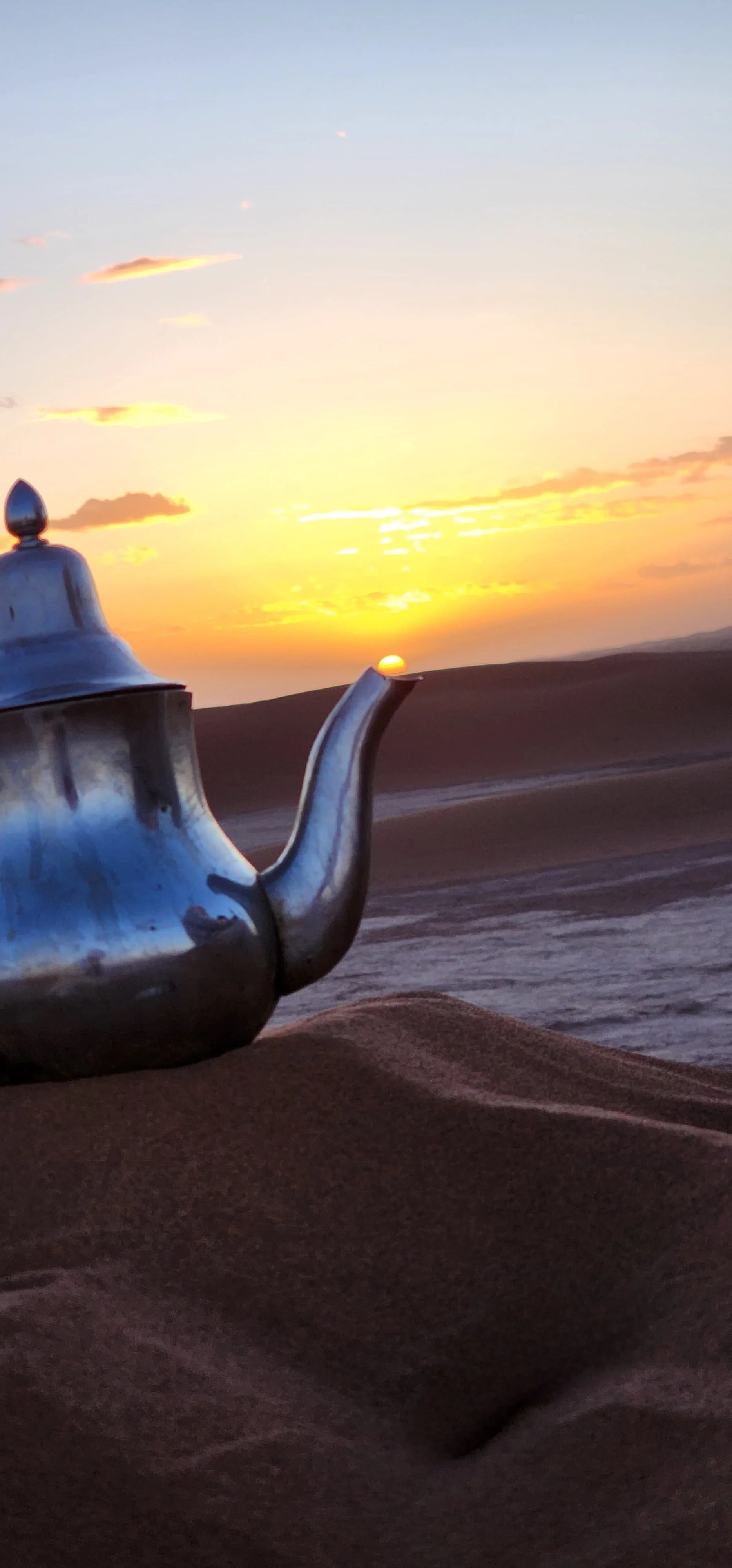 Une théière en métal argenté posée sur du sable dans un désert avec un coucher de soleil à l'horizon.