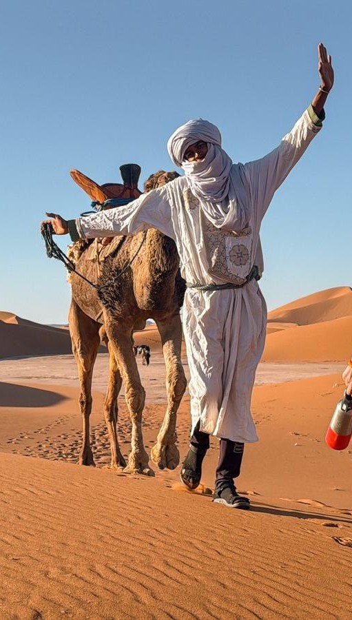 Un chamelier en tenue traditionnelle marocaine dans le désert, avec un dromadaire derrière lui, le tout sous un ciel bleu clair.