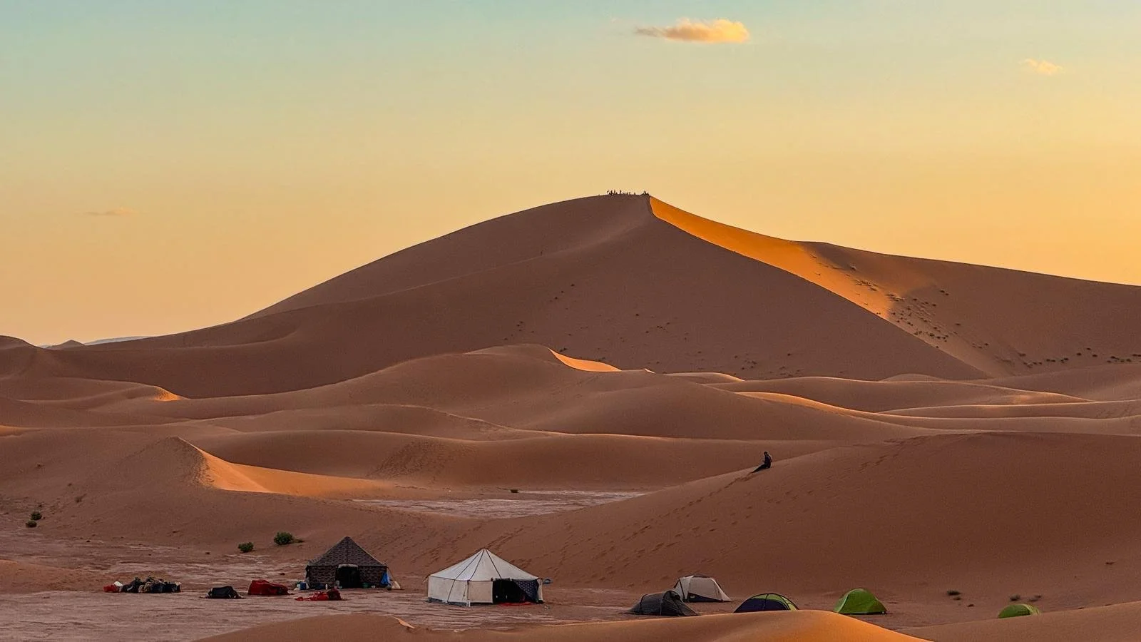 Camping dans le désert avec des tentes et une grande dune de sable au coucher du soleil.