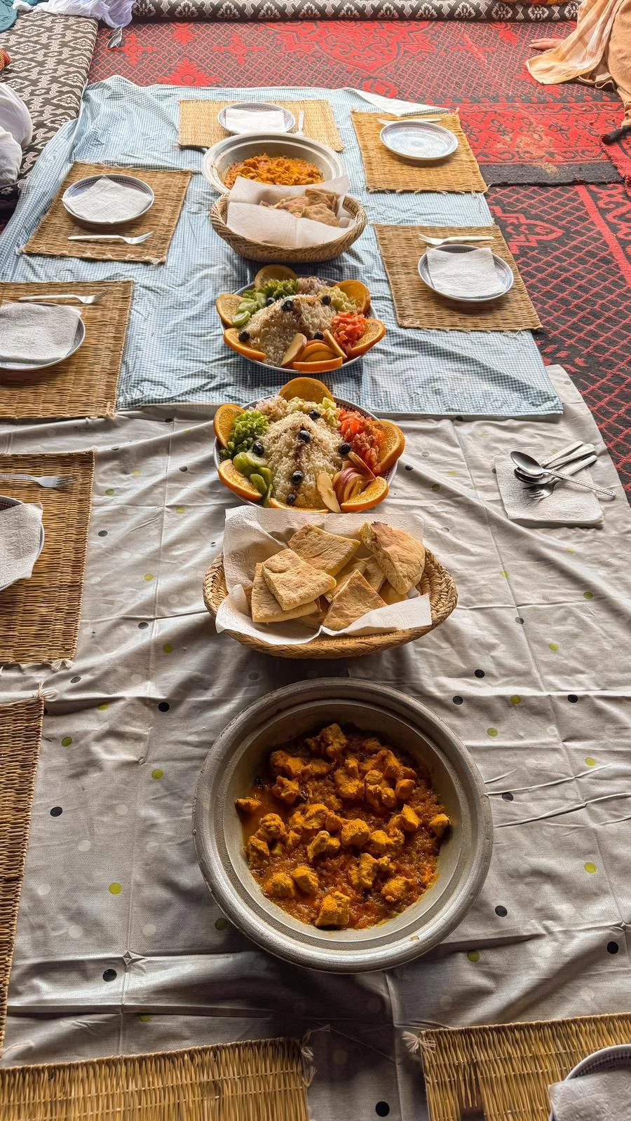Repas traditionnel africain avec plats en sauce, riz, pain plat, fruits et légumes, posé sur une nappe à carreaux et une nappe blanche, avec des couverts en métal et des assiettes en plastique.