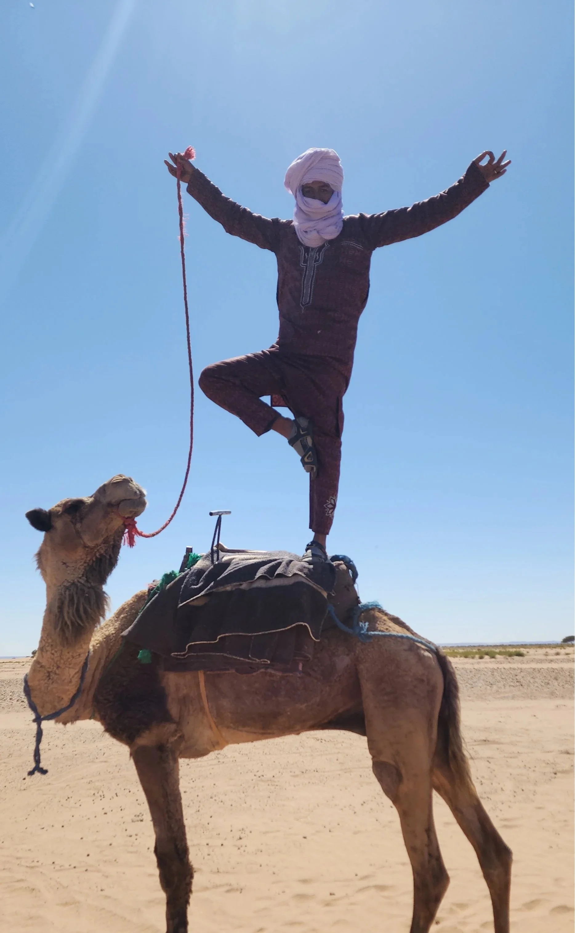 Un homme habillé en vêtements traditionnels marocains et un chèche blanc noué sur sa tête se tient debout sur un chameau dans un désert, avec un ciel bleu clair.