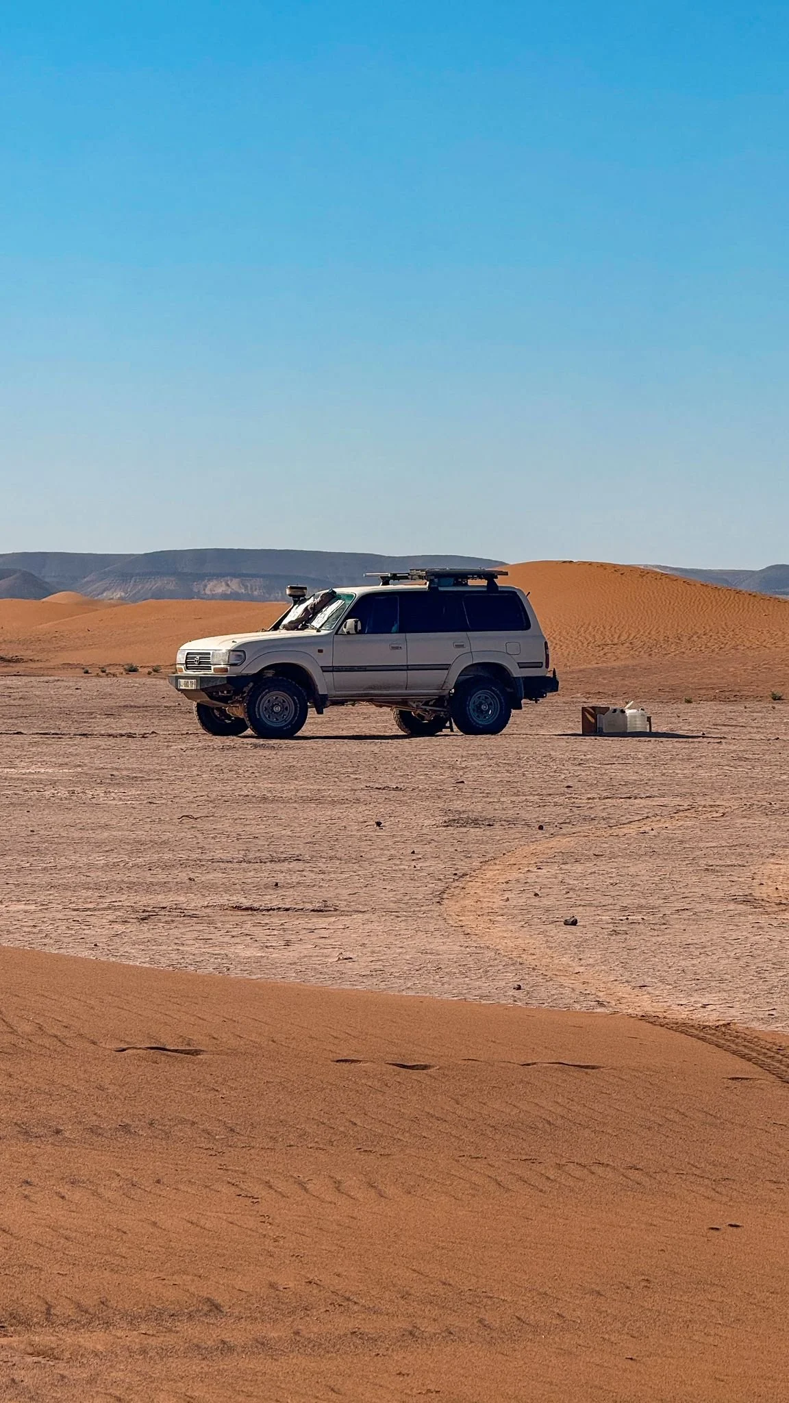 Voiture blanche dans un désert avec dunes de sable et horizon lointain, ciel bleu clair.