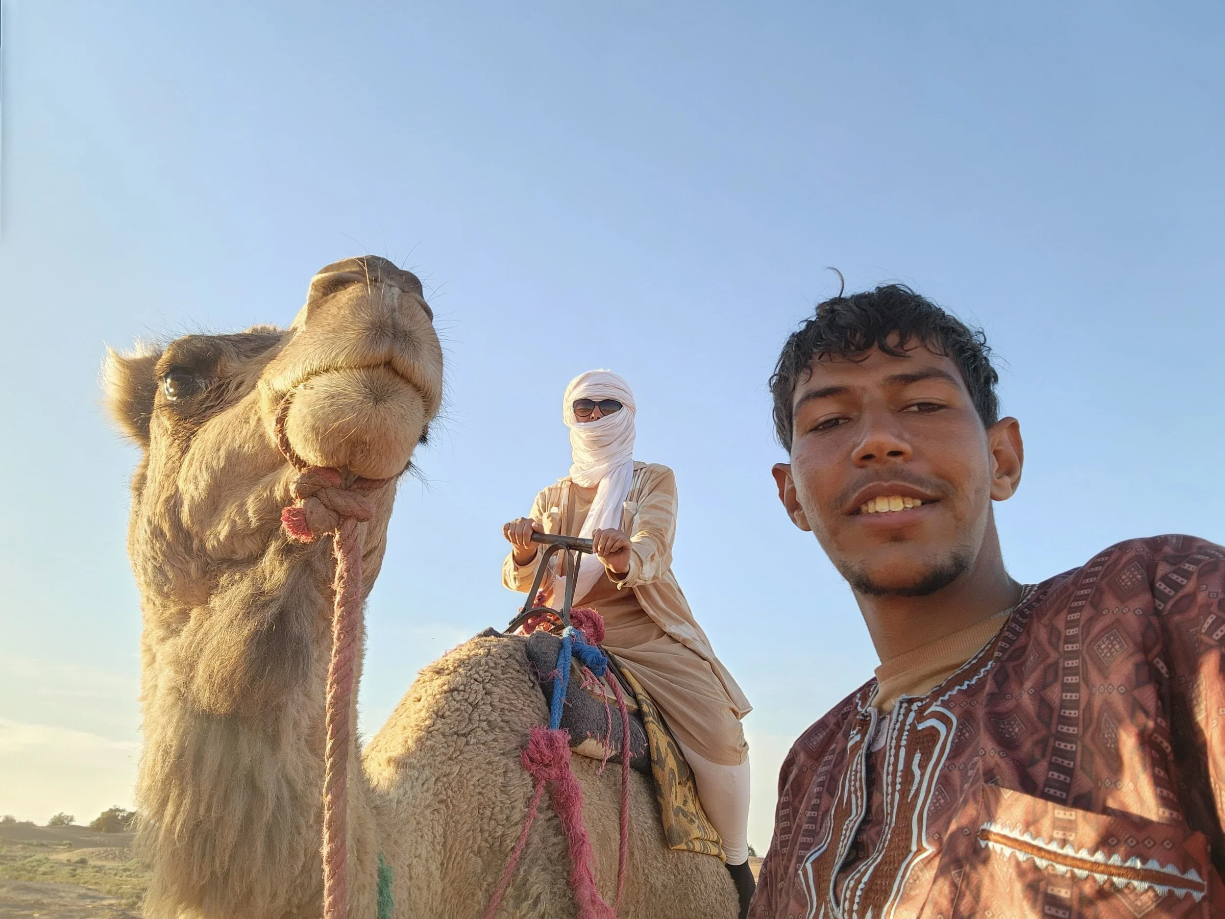Une touriste française portant un chèche nouée sur sa tête et des lunettes de soleil est sur un dromadaire guidé par un nomade marocain sous un ciel clair. 