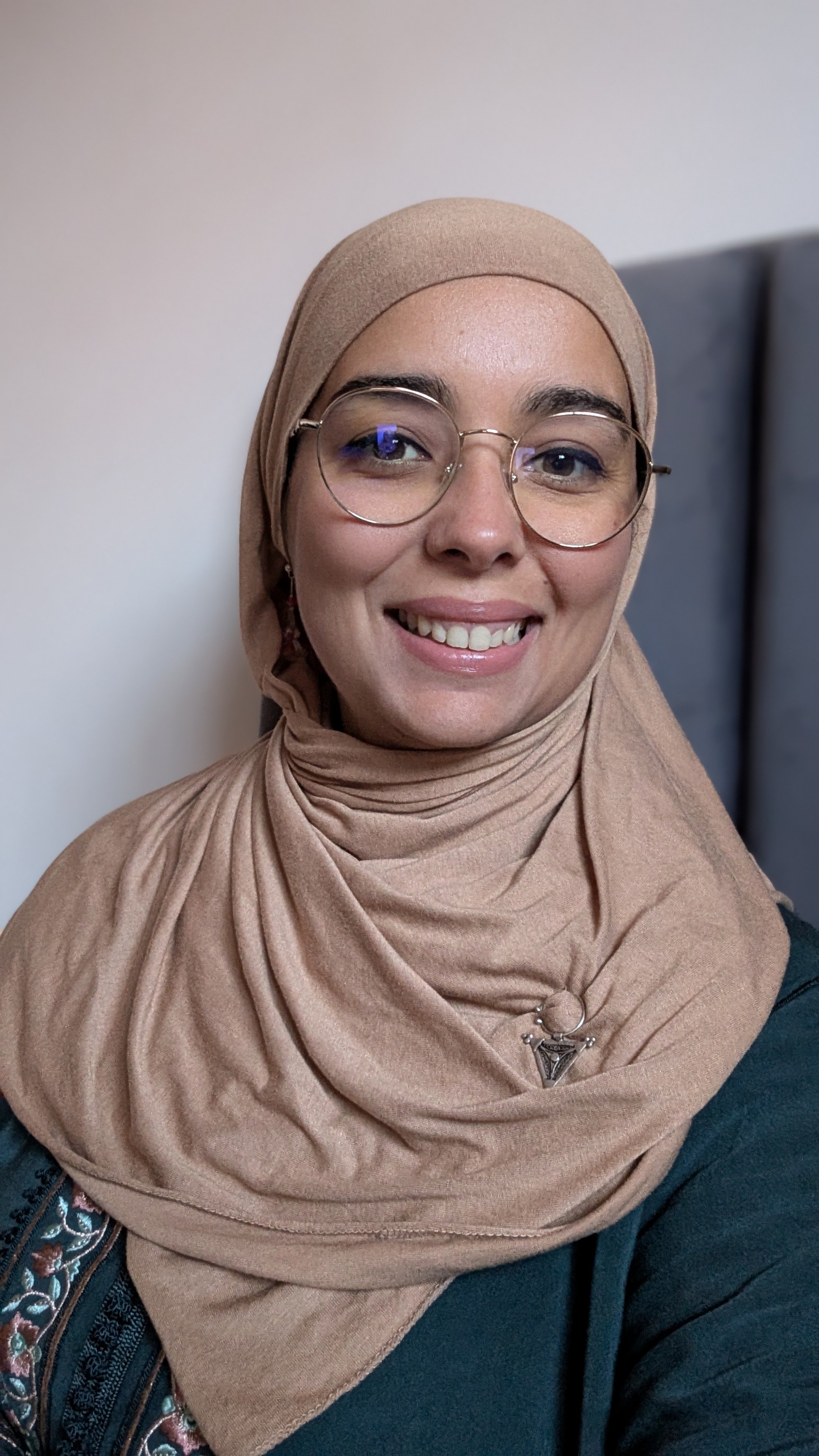 Portrait d'une femme souriante portant un hijab beige et des lunettes rondes, avec un fond neutre.