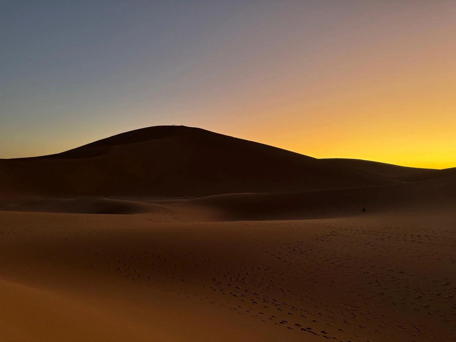 Sable chaud du désert avec une grande dune, le coucher du soleil à l'horizon, ciel dégradé allant du bleu au jaune orangé.