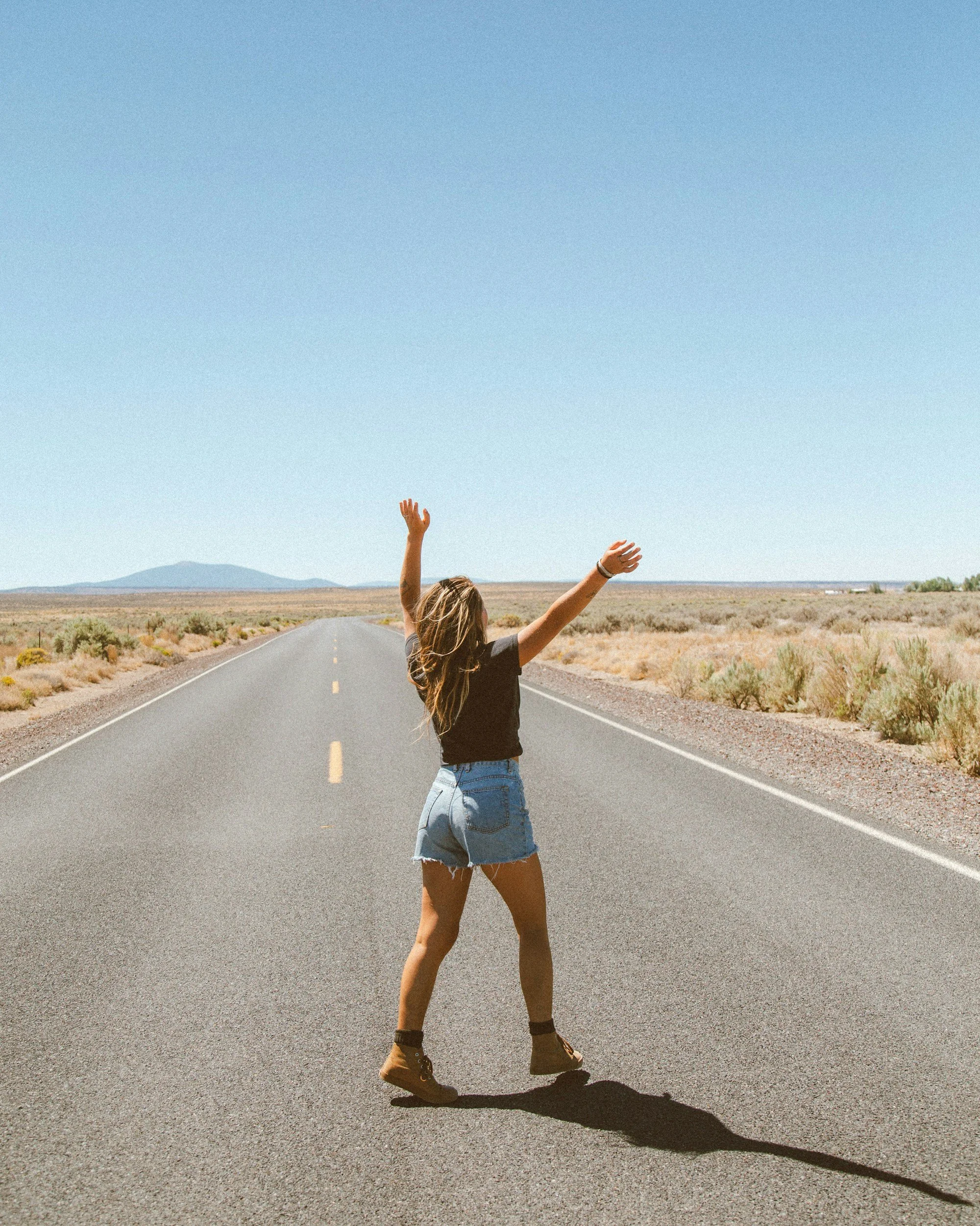A person with long hair, wearing a black t-shirt, denim shorts, and brown boots, is standing on an empty paved road in a desert landscape. They are raising their arms with hands open and facing upward, and casting a long shadow on the ground against a clear blue sky.