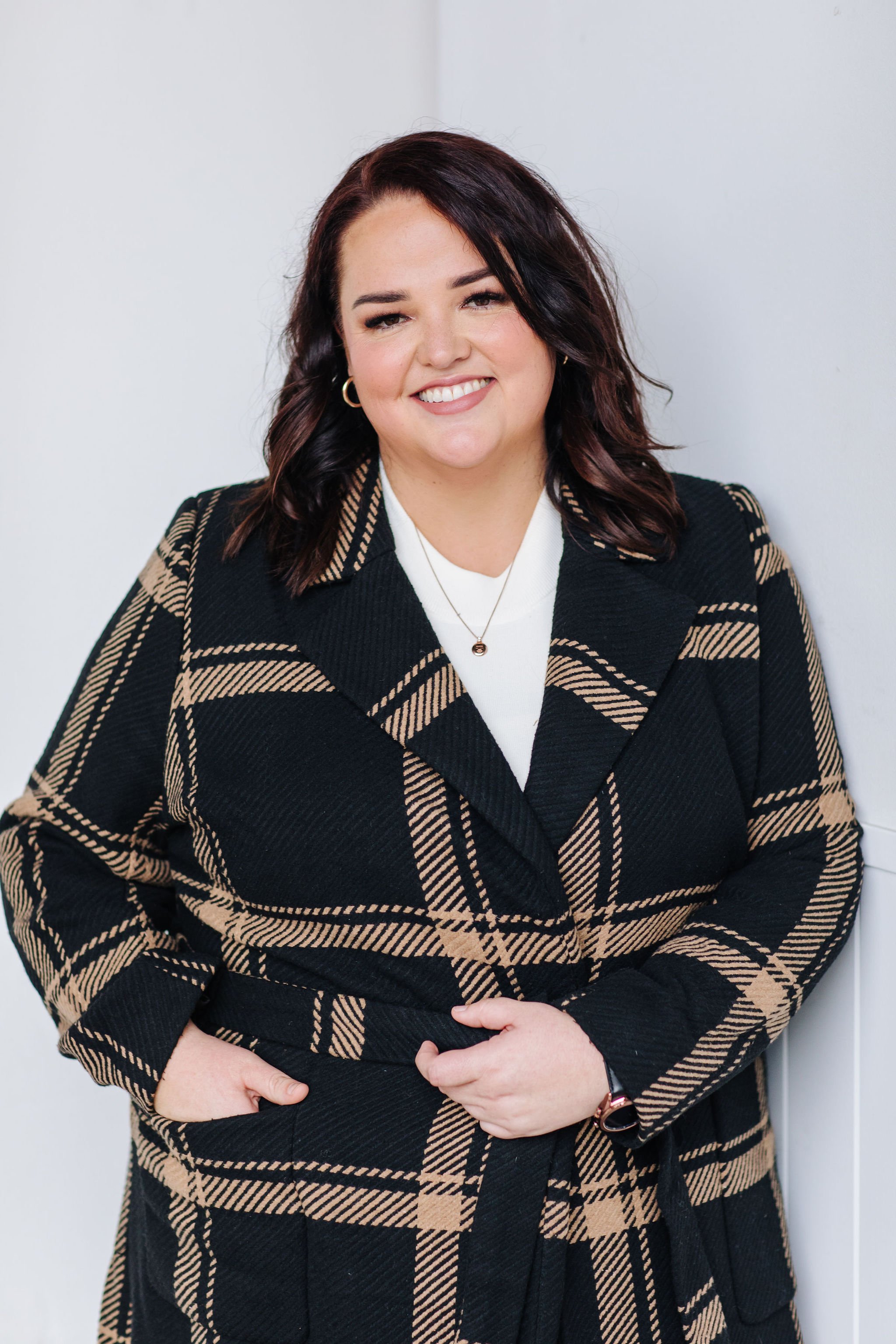 A woman with dark wavy hair smiling, wearing hoop earrings, a white top, a necklace, and a black and beige plaid coat, standing against a plain white wall.