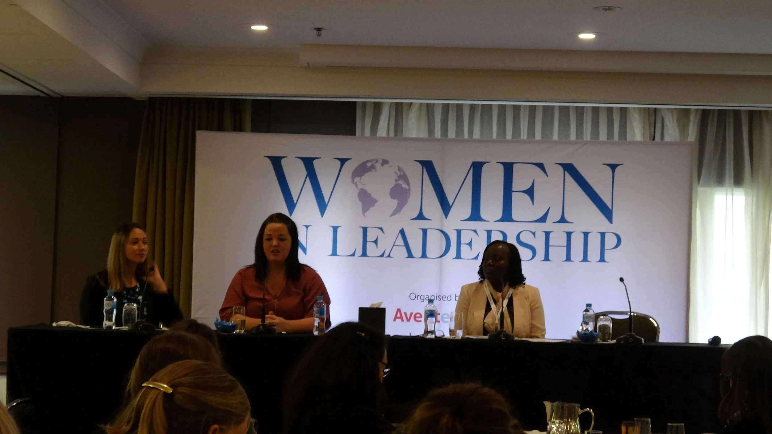 Three women sitting at a panel during a women leadership conference, with a large banner behind them that reads 'Women in Leadership' and a globe graphic inside the 'O' in 'Women.'
