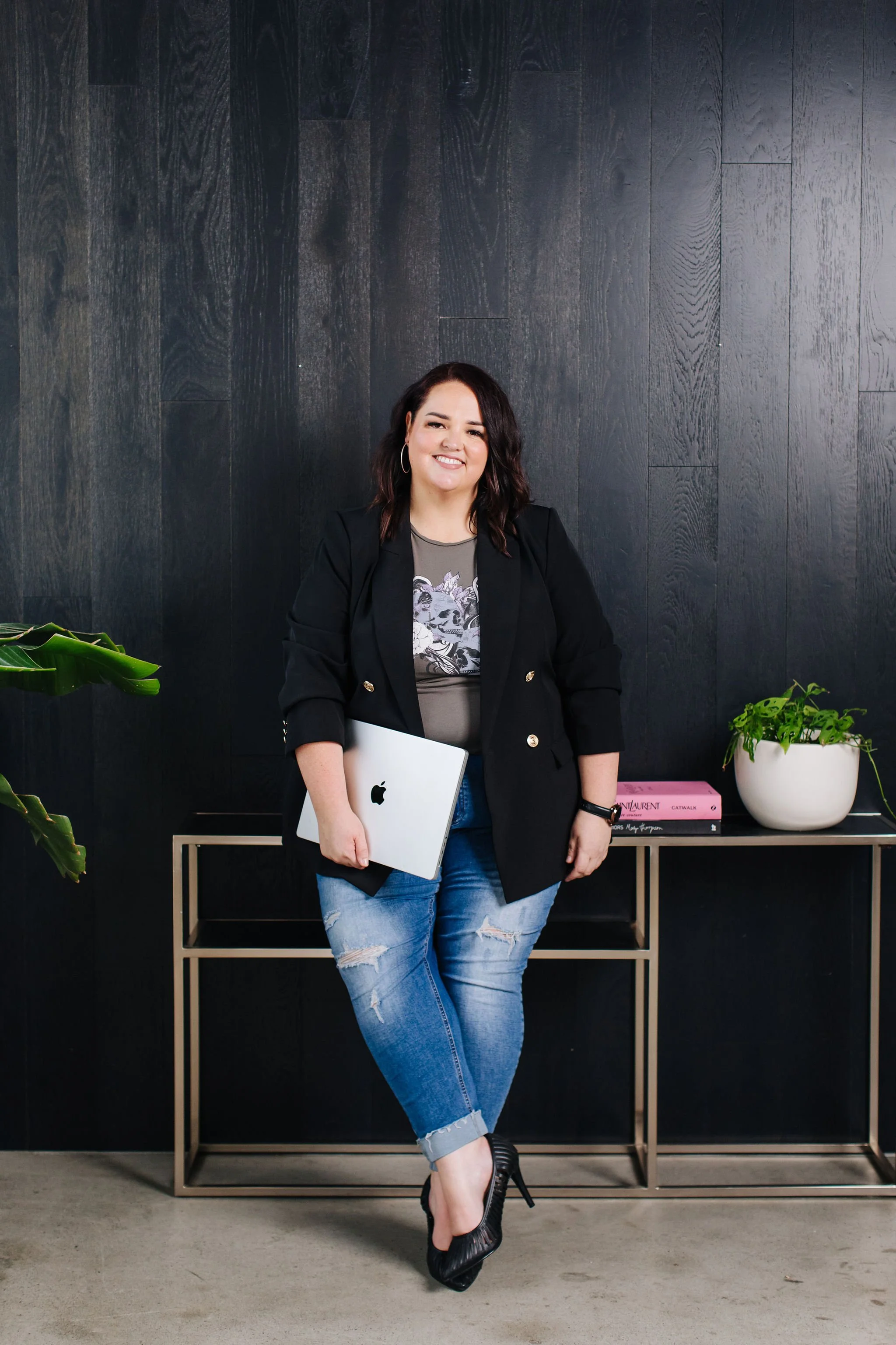 A woman standing inside an office, holding a silver MacBook, wearing a black blazer, gray graphic t-shirt, ripped blue jeans, and black high heels, in front of a black wooden wall with a beige table, pink and black books, and green plants.